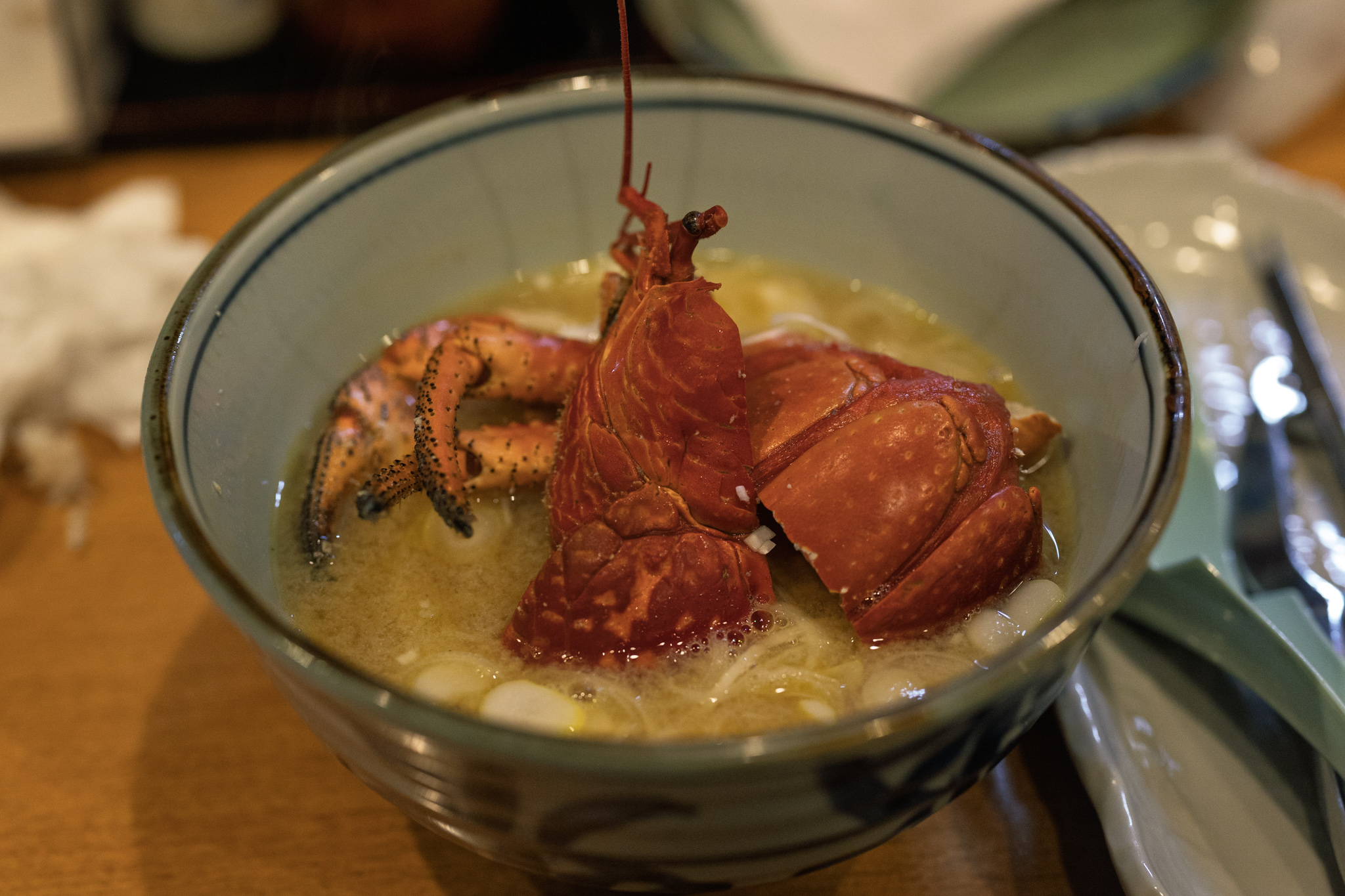 A bowl of coconut crab soup with the red shell split and standing upright, legs folded beside it, and a long antenna and stalked eyes rising above a cloudy broth