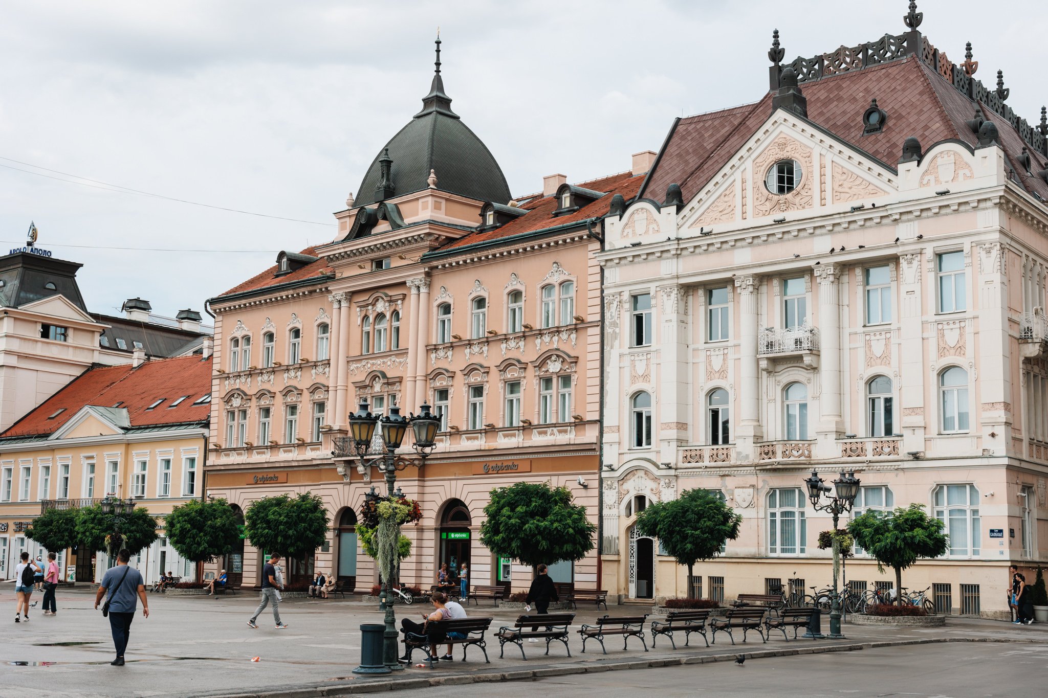 Ornate pastel-coloured buildings around Freedom Square in Novi Sad, with people walking and sitting on benches.