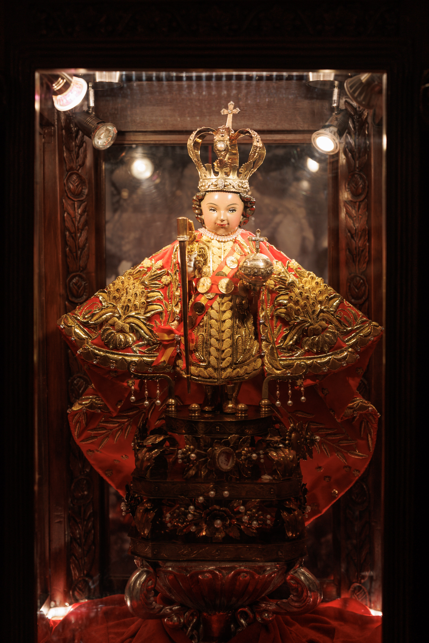 A small statue of the Child Jesus in a tall gold crown and a red cape heavily embroidered in gold, holding a gold sceptre and an orb topped with a cross, a sash of coins across his chest, in a lit glass case