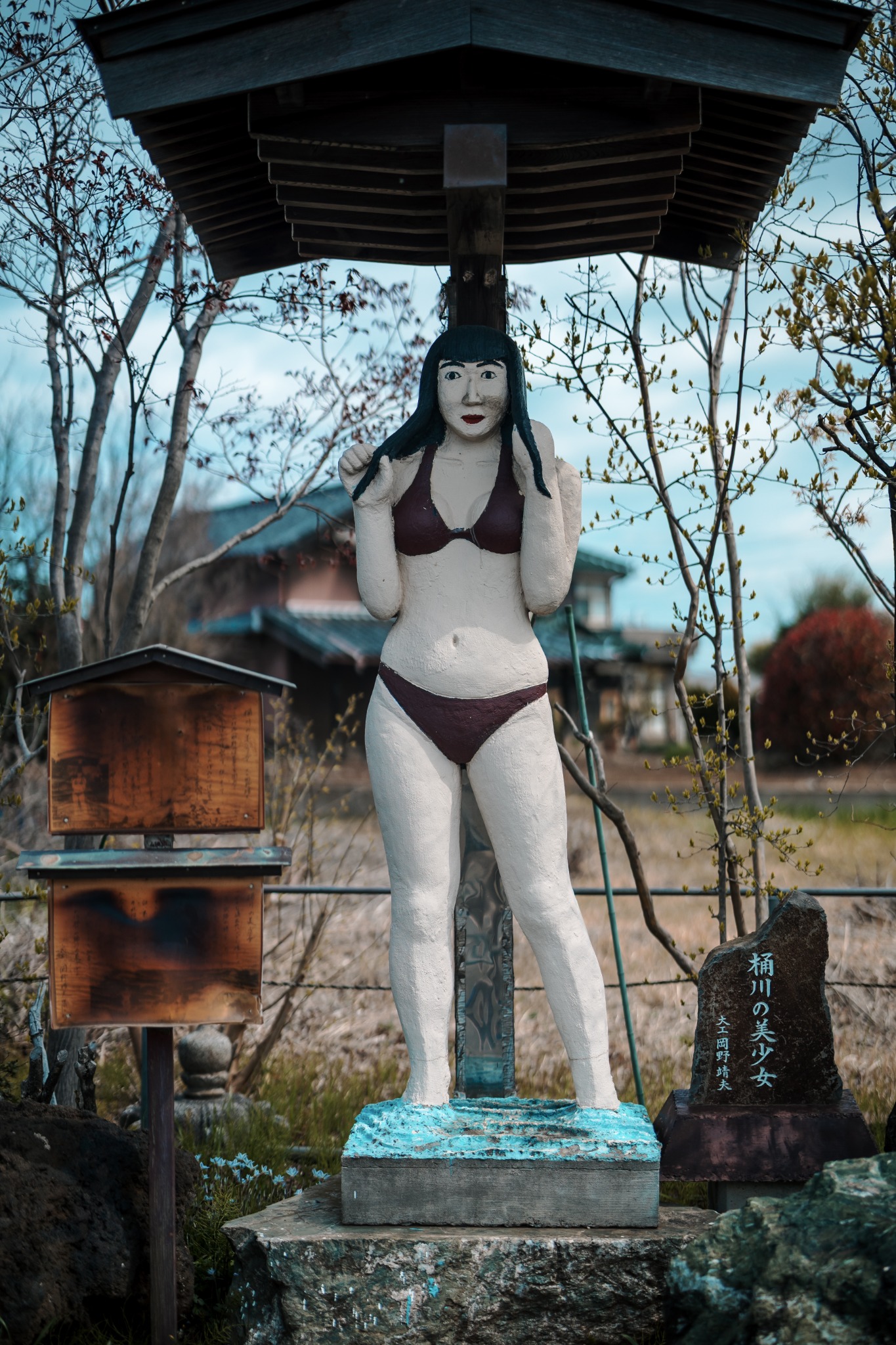 Full-length concrete figure of the Beautiful Girl of Okegawa in a swimsuit, standing beside a stone plaque bearing her name in Saitama.
