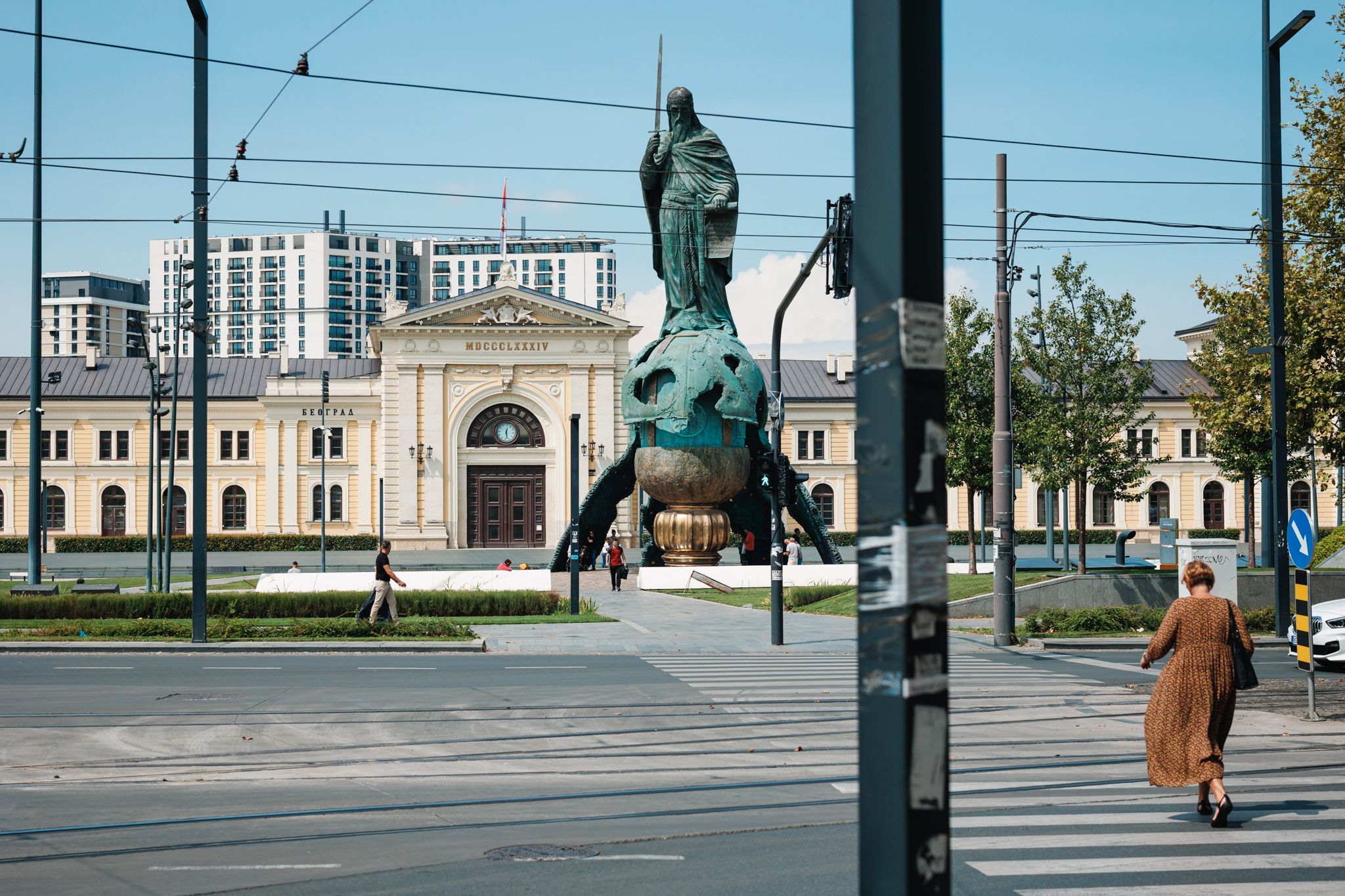 Belgrade’s former main railway station and the Stefan Nemanja monument across the tram tracks on Savski Square, with pedestrians in the foreground.