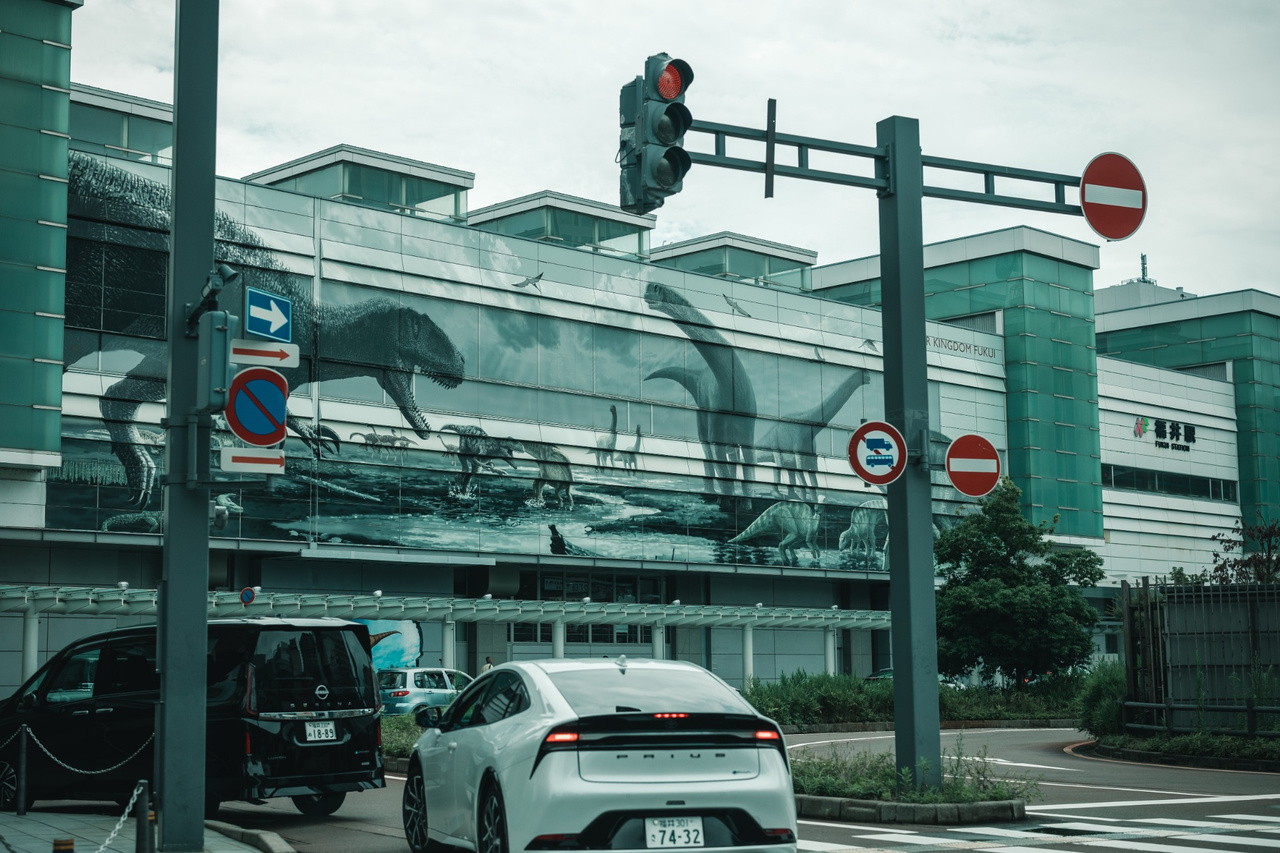 Large dinosaur mural covering the west-side wall of Fukui Station, seen across the station forecourt and road traffic.