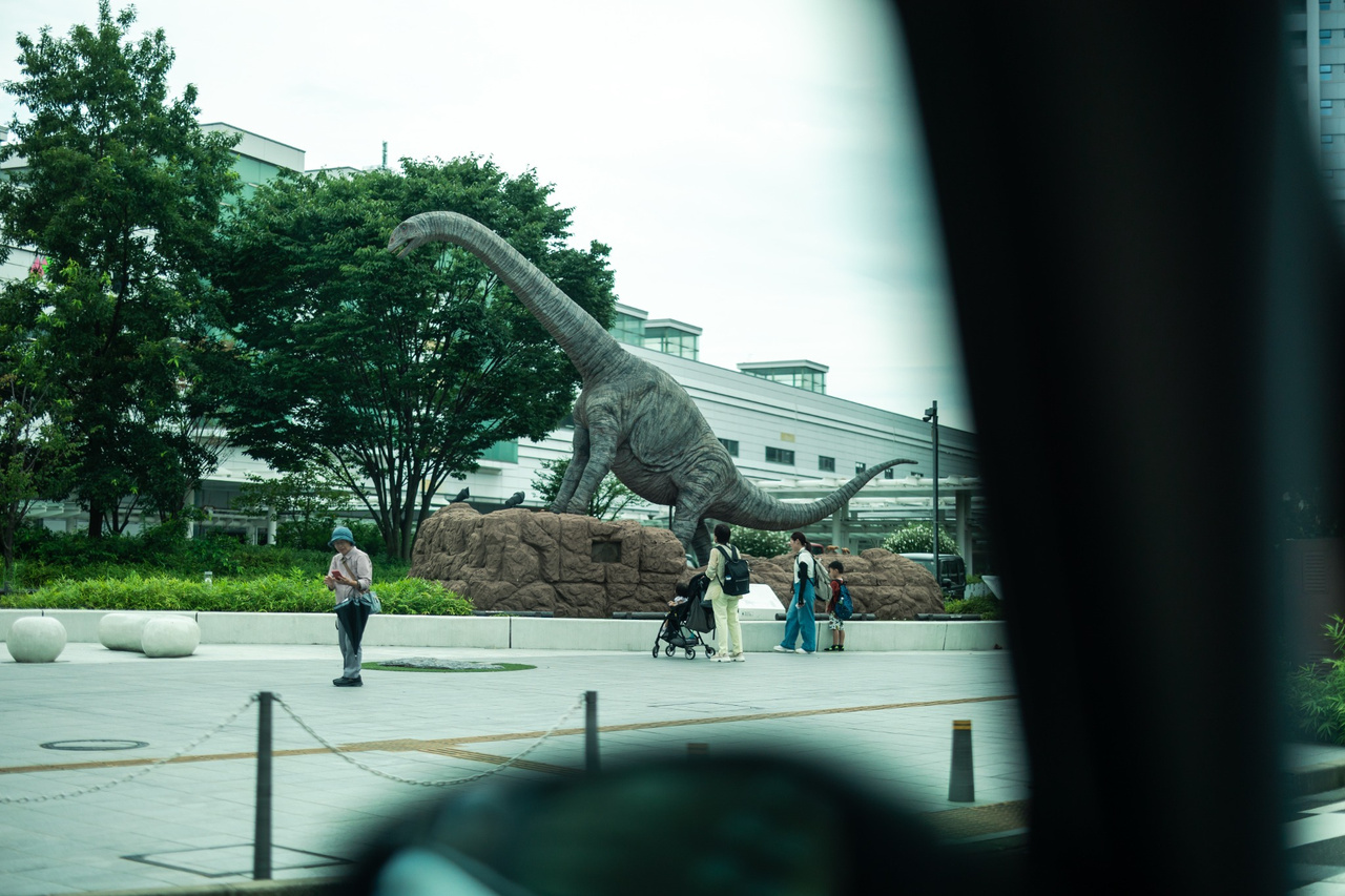 Full-scale Fukuititan animatronic in the Dinosaur Plaza outside Fukui Station, with people nearby.