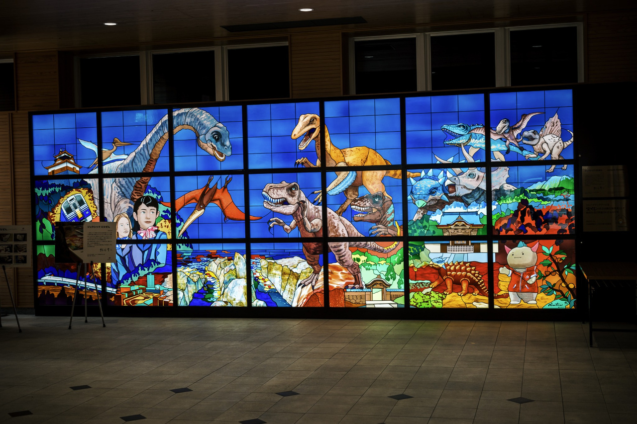 Large illuminated Jurassic Echizen stained-glass artwork filled with dinosaurs and Fukui landmarks at Echizen Railway Fukui Station.