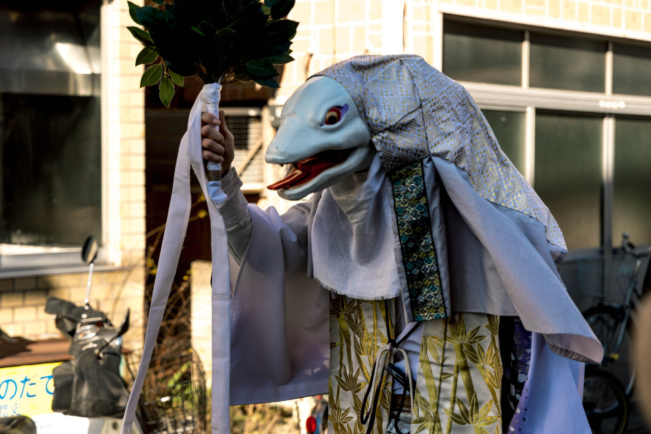 The white-snake performer holding a leafy branch, with neighbourhood buildings behind.