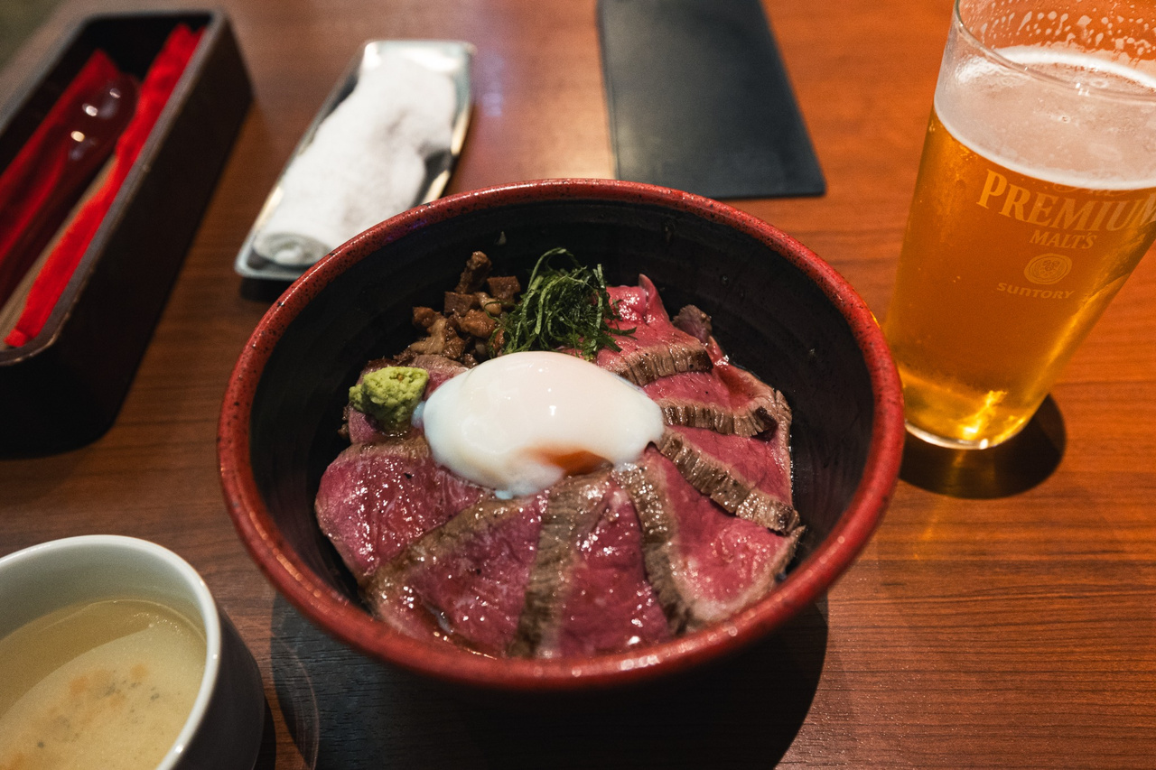 Akaushi don on a table at yoka-yoka in Kumamoto, with an onsen egg, wasabi, a beer glass and a soup bowl nearby.