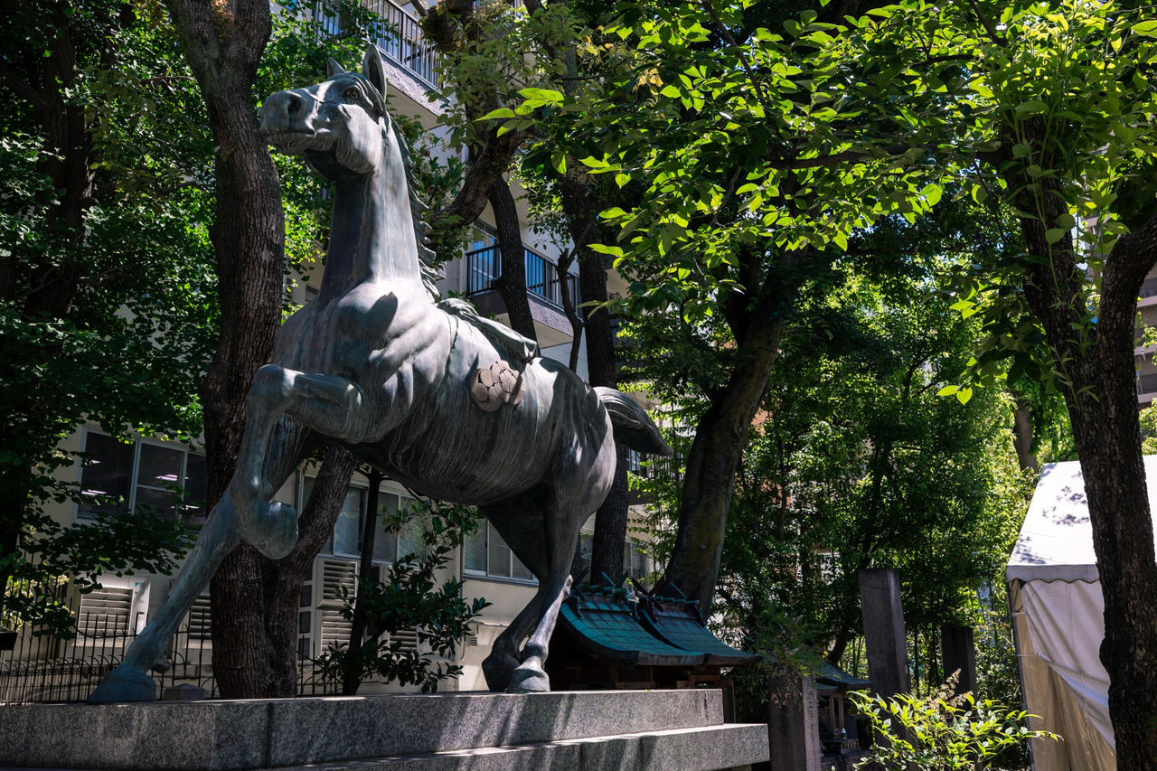 Bronze statue of a sacred horse in mid-stride, one foreleg raised, on a stone plinth among trees at Namba Yasaka Shrine.