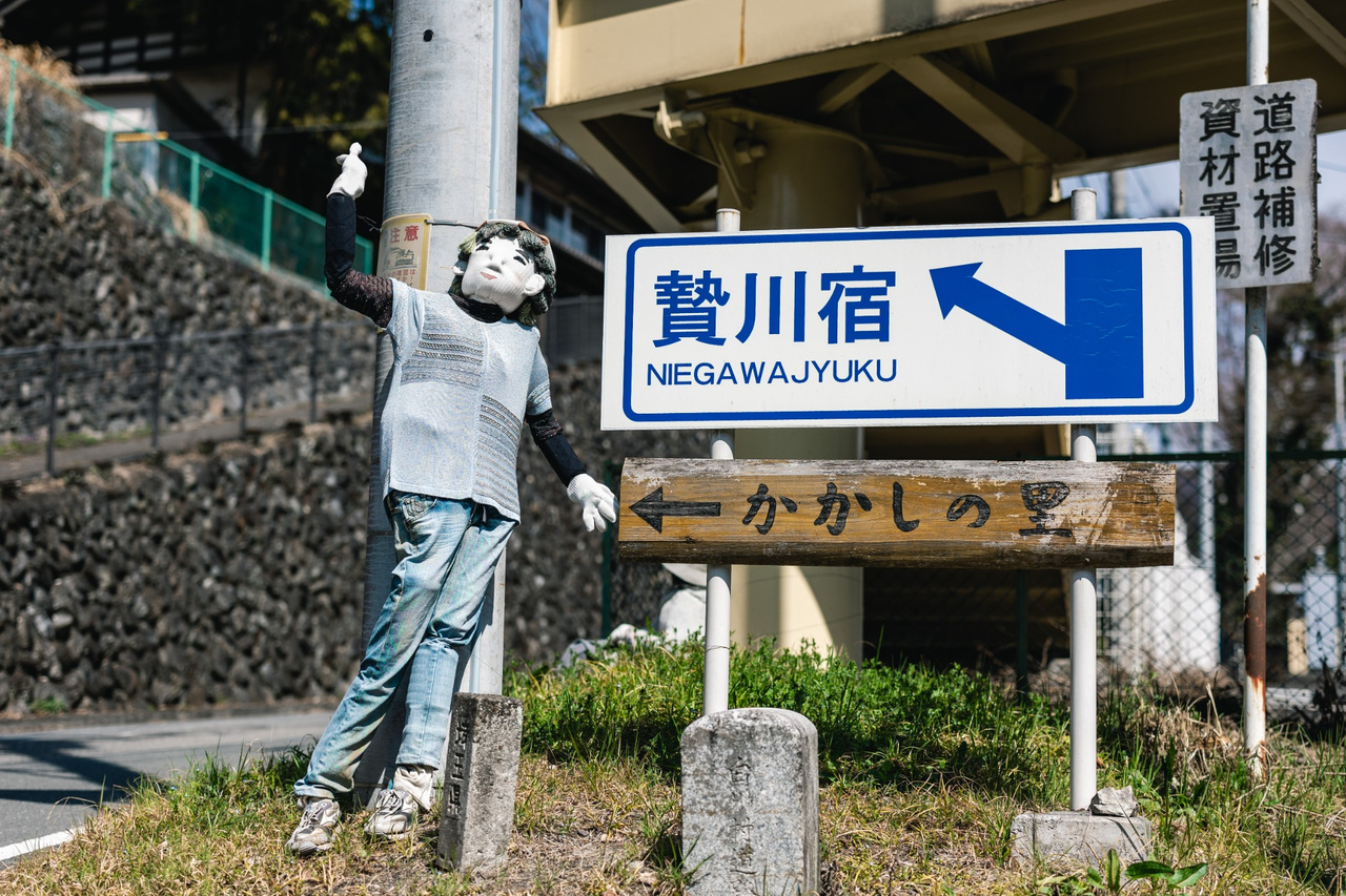 A handmade scarecrow raises a gloved hand beside the Niegawa-juku sign in Chichibu.