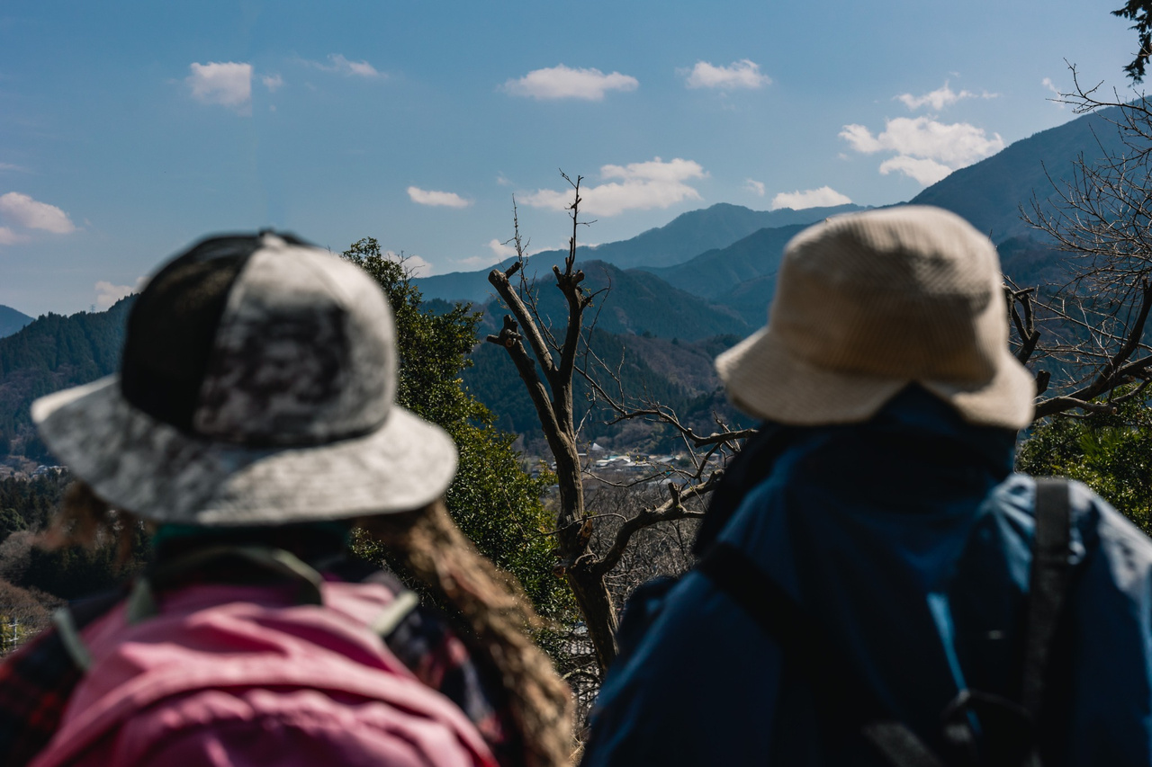 Two handmade scarecrows wearing hats and backpacks face mountains beyond Niegawa-juku.