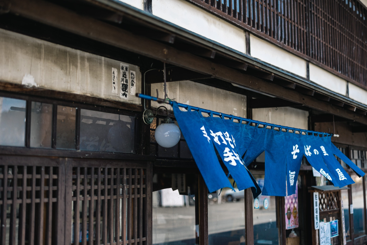 Blue noren curtain at the entrance to Fukushimaya, marked with handwritten Japanese for handmade soba and udon.