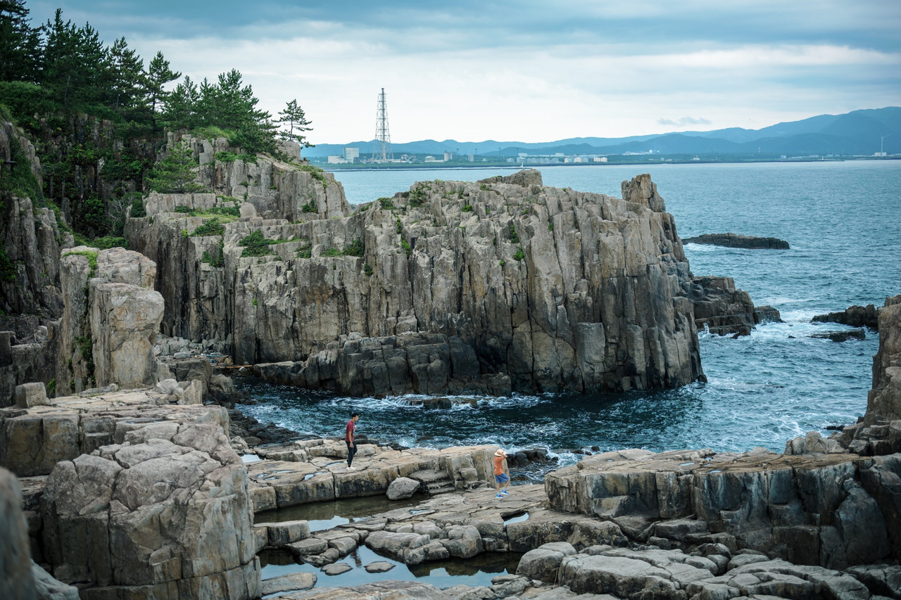 Tall columnar cliffs topped with pine trees at Tojinbo, with a steel lattice tower and industrial buildings across the water.