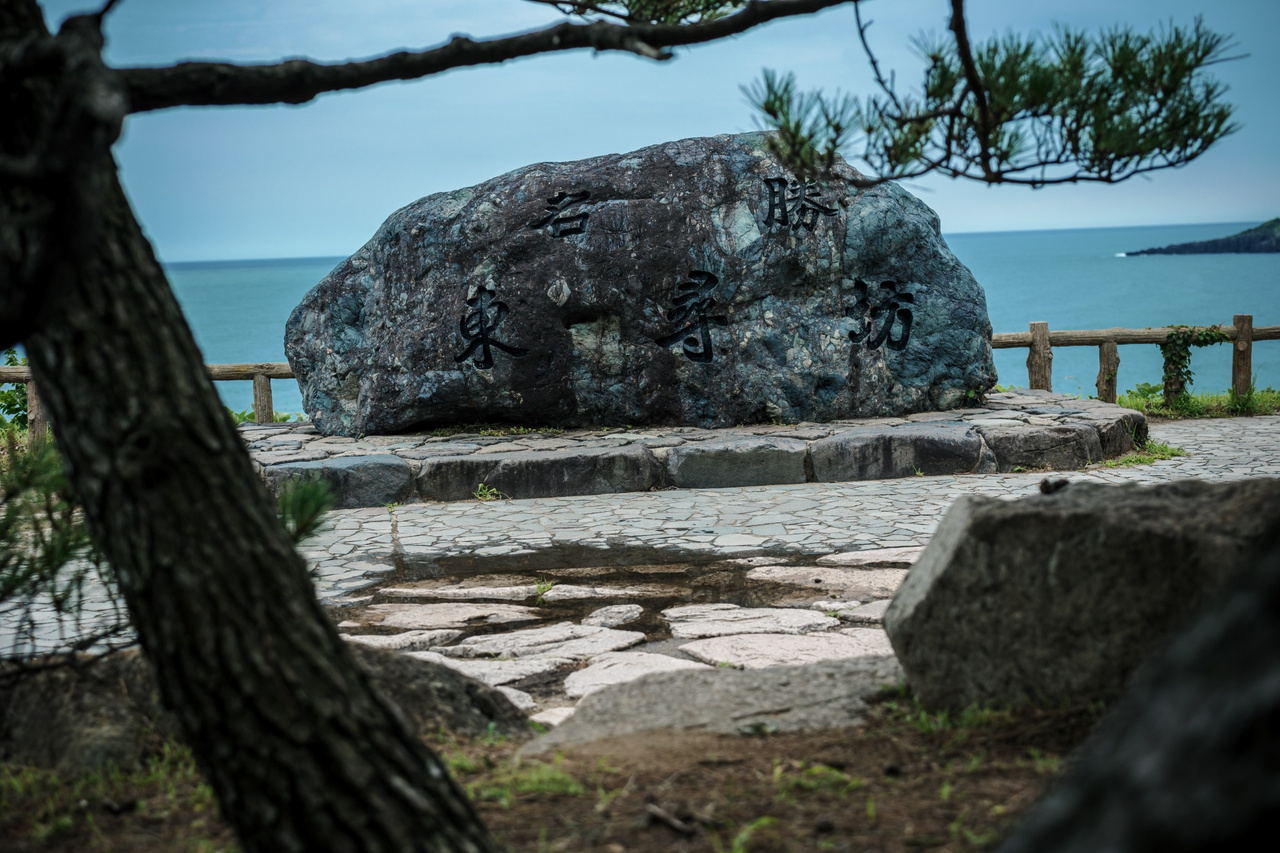Large boulder inscribed in Japanese with “Place of Scenic Beauty: Tojinbo,” seen past a pine branch with the Sea of Japan behind.