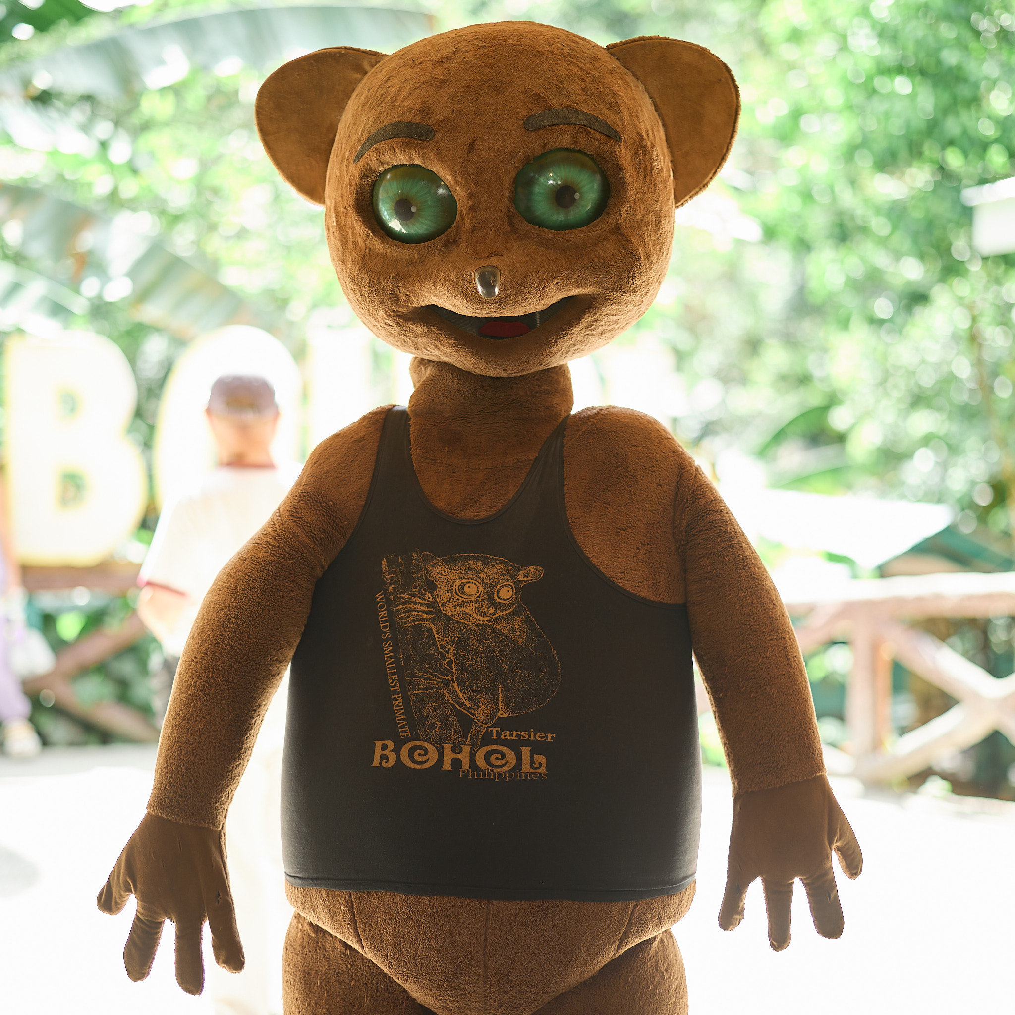 The tarsier mascot facing the camera head-on, with large green eyes, a small nose and a smiling mouth, its black shirt printed with a drawing of a tarsier and the words WORLD’S SMALLEST PRIMATE, Tarsier, BOHOL, Philippines