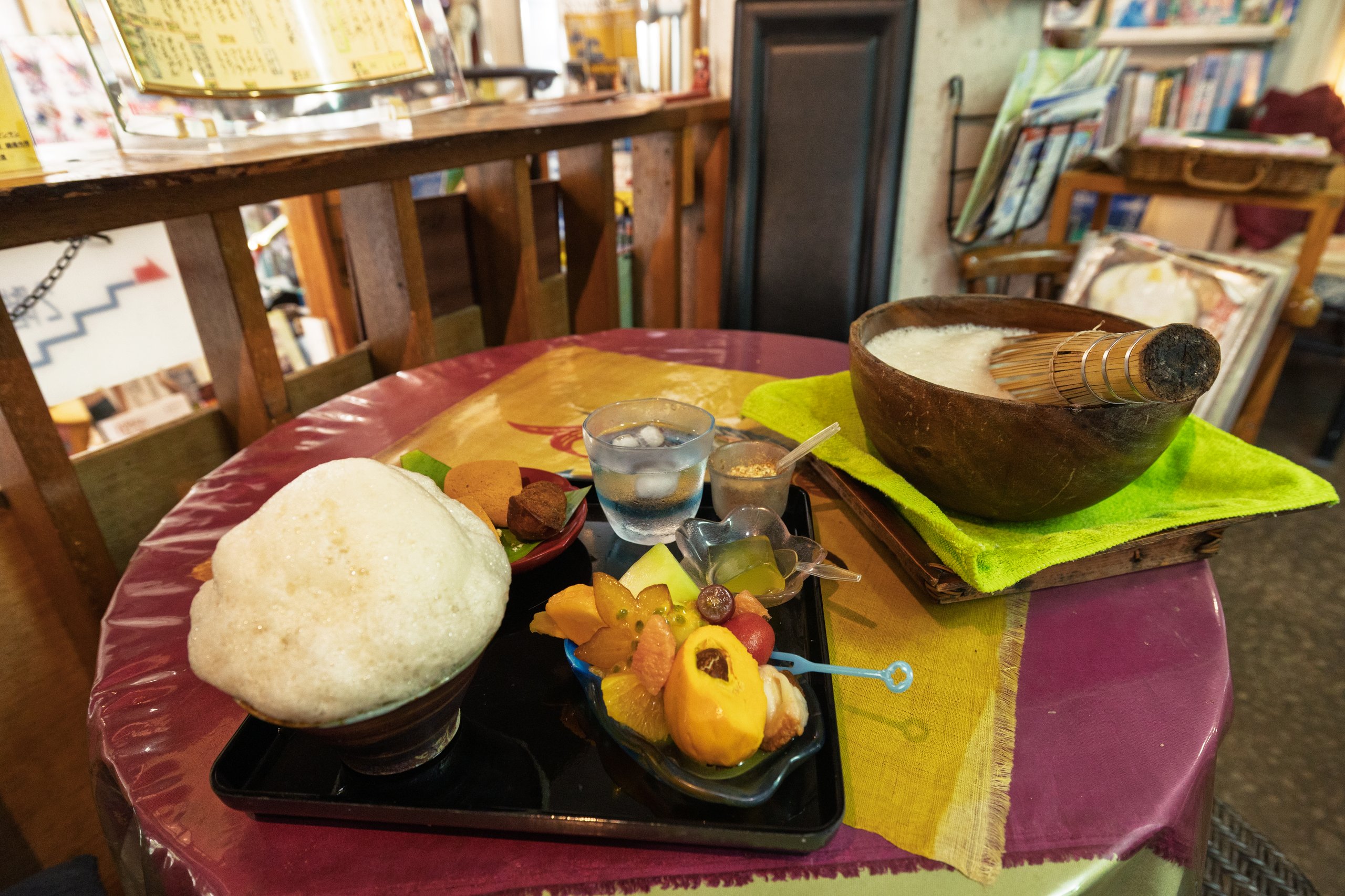 A bukubuku tea set on a black tray: a cup piled high with white foam, small brown sweets, iced water, a cup of brown sugar and peanuts, a little dish of green jelly and a plate of tropical fruit, with a large wooden bowl and bamboo whisk on a green cloth beside it