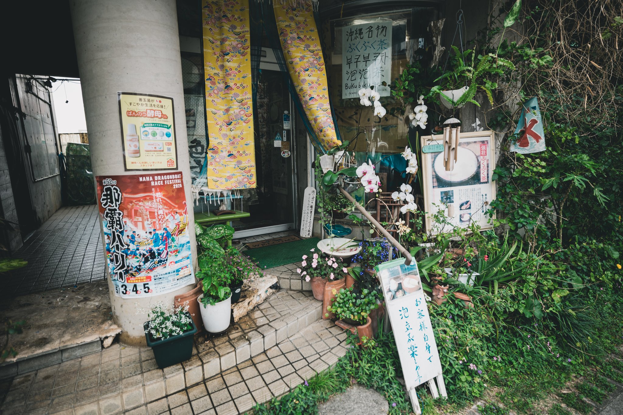 The entrance of a small tea house crowded with potted plants and white orchids, with patterned yellow curtains, posters and a hand-painted white board in Japanese at the bottom right