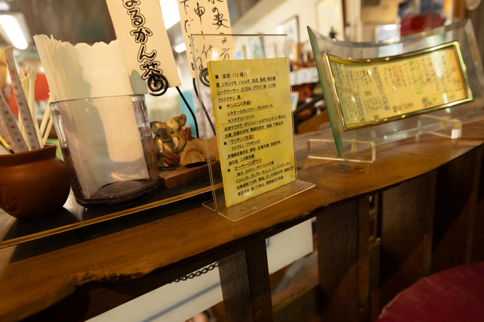 A yellow card in a clear stand on a wooden counter listing tea bases in Japanese — thirteen herbs, shell ginger, turmeric and bitter melon — beside a glass of straws and paper napkins