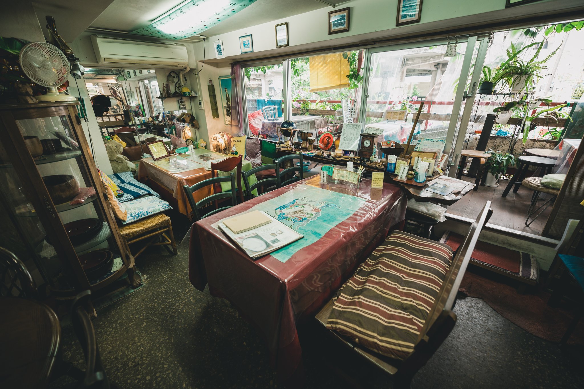 The seating area of the tea house: tables with red covers, mismatched chairs and cushions, a glass cabinet of bowls, and plants filling the windows