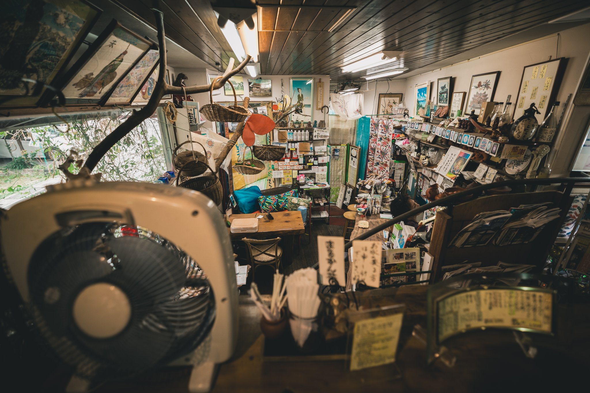 Looking down into the tea house past an electric fan: a low counter, shelves of goods, framed pictures, baskets and a wall covered with small items
