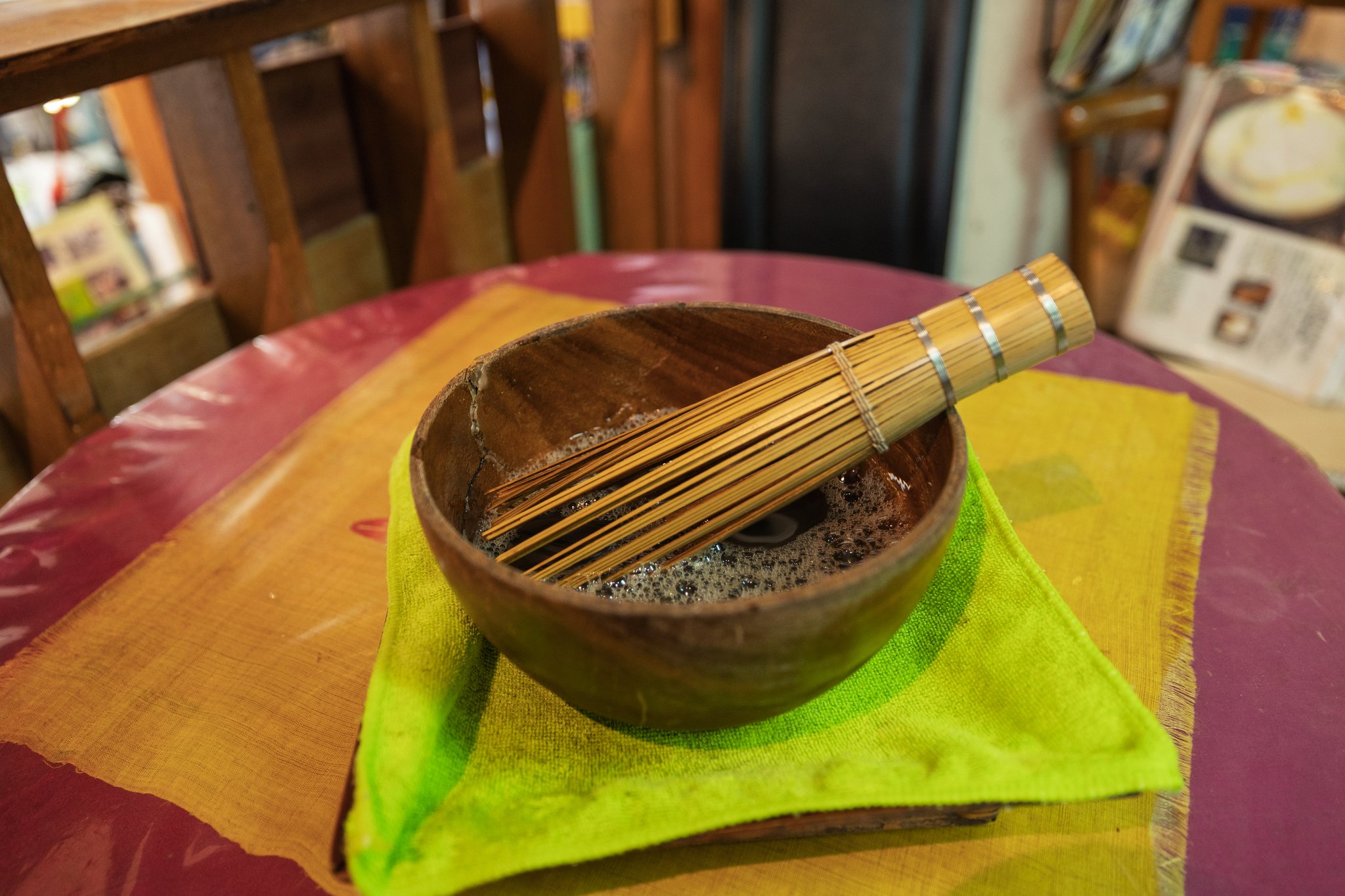 A wooden bowl of brown liquid with only a few bubbles, a long bamboo whisk resting across it, on a bright green cloth