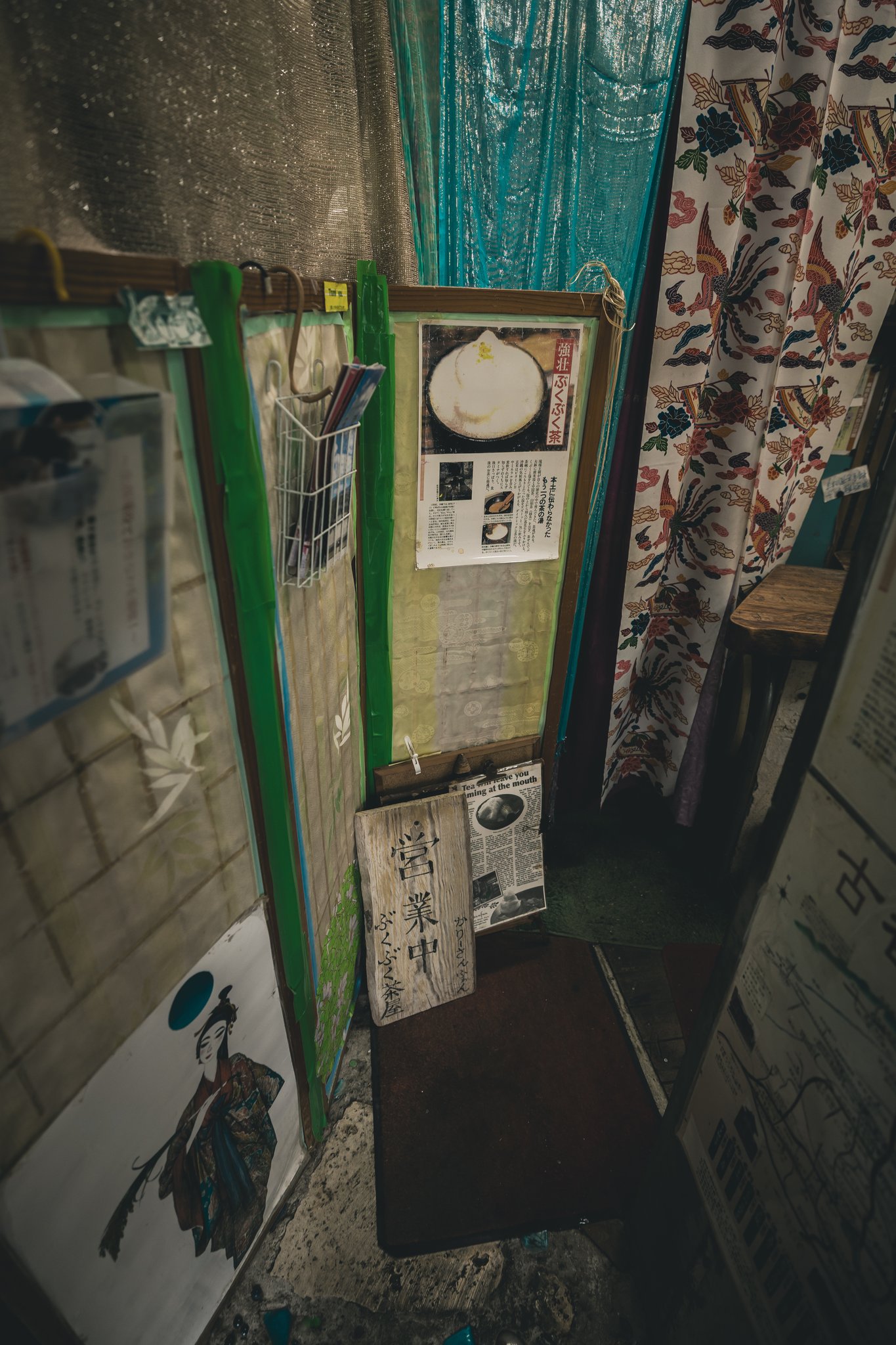 A weathered wooden sign reading 営業中 and ぶくぶく茶屋 in a narrow entranceway, leaning against an English newspaper clipping headlined “Tea will leave you foaming at the mouth”, below a poster of a bowl of foam