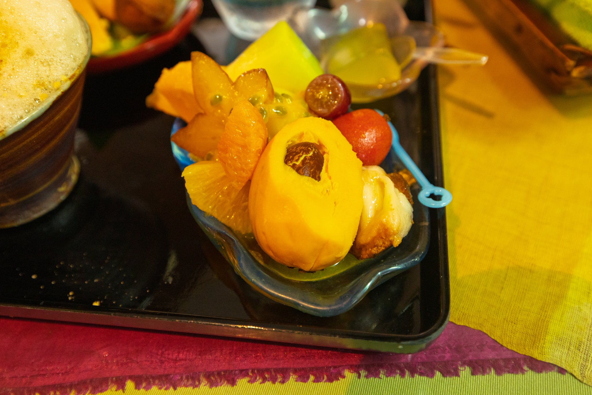 A blue glass dish of tropical fruit: a halved yellow canistel with a large brown seed at the front, star fruit and passion fruit pulp at the back, citrus segments, a grape, a red acerola and a white piece of unidentified fruit, beside a cup of foam