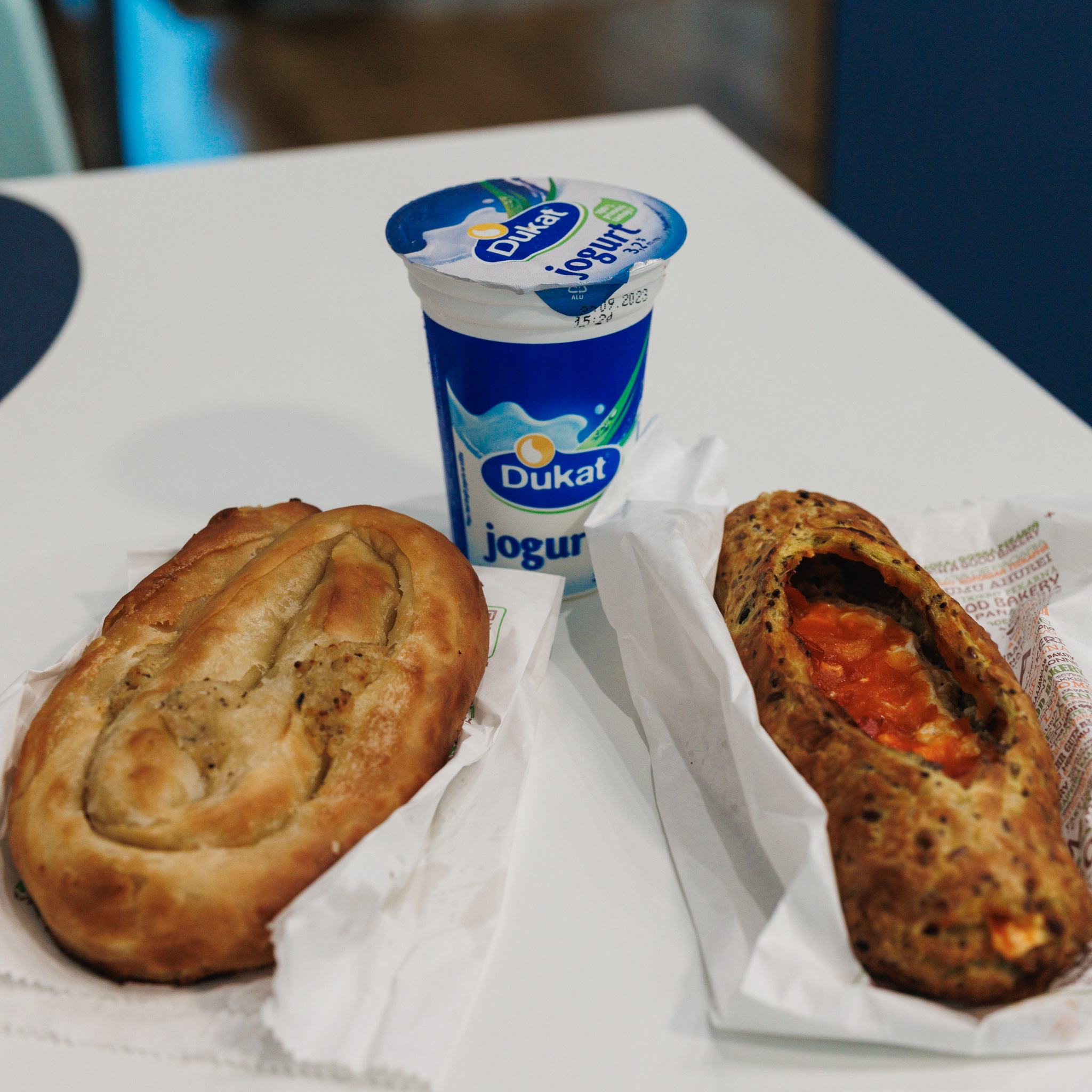 A meat-filled burek and an ajvar-and-cheese pastry on white paper beside a cup of yogurt at Skroz Dobra Pekara in Novi Sad.