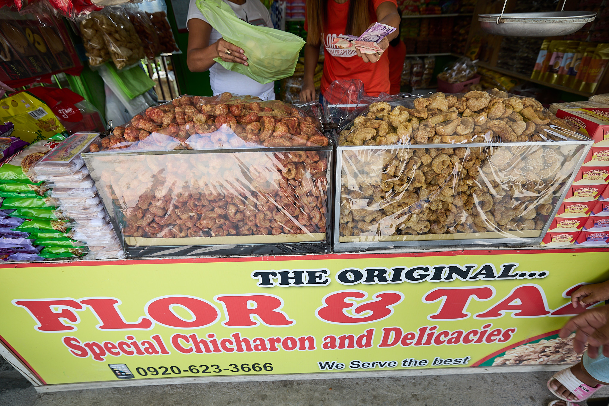 Two glass cases heaped with chicharon at the FLOR & TAZAR stall in Carcar, one pile reddish brown and the other pale golden, with a hanging scale above and a hand holding out peso notes.