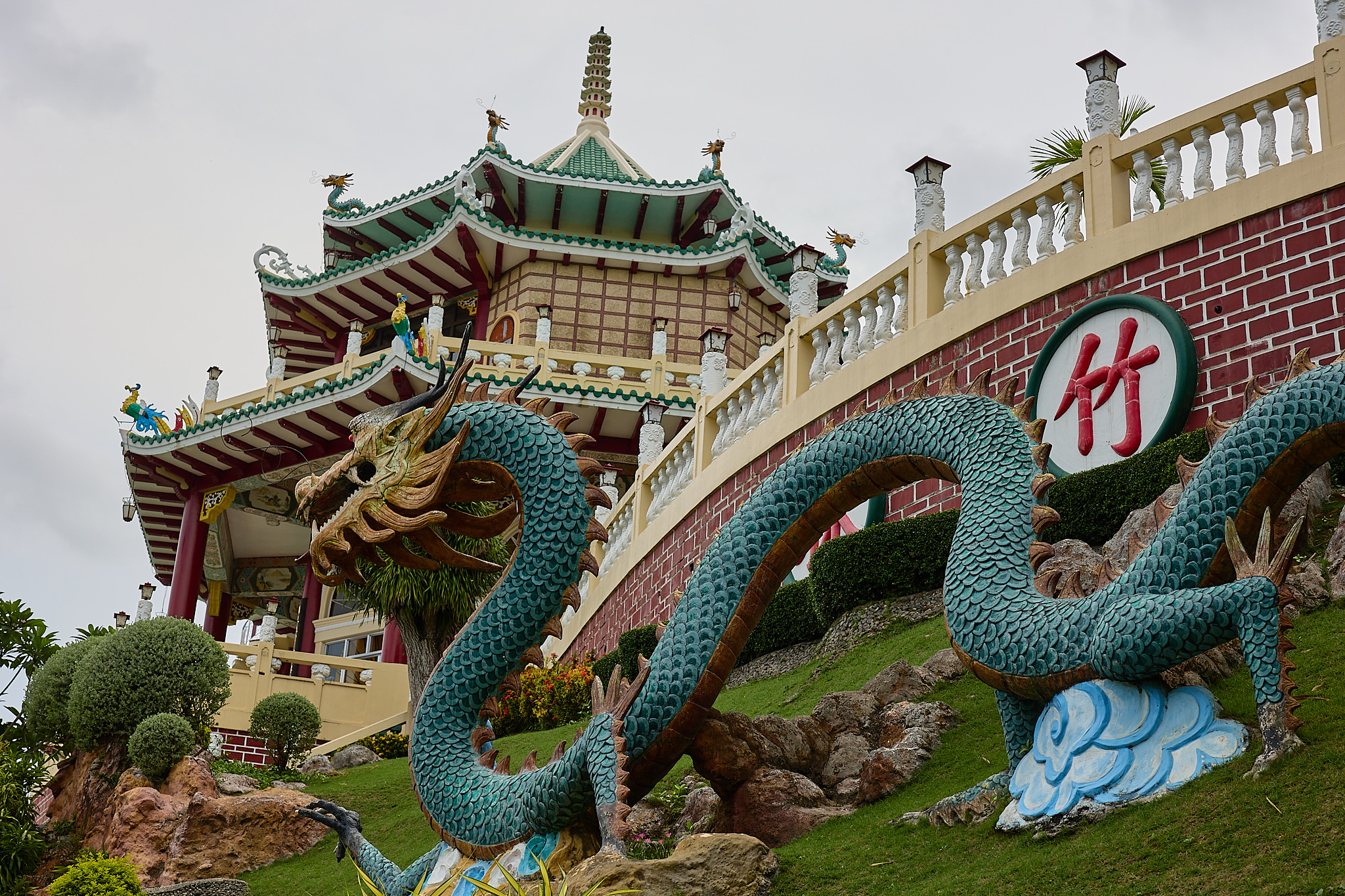 A sculpted dragon with a golden head and blue-green scales winding up a grassy slope towards a multi-tiered pavilion with green roofs, beside a brick wall with a round panel bearing a red Chinese character