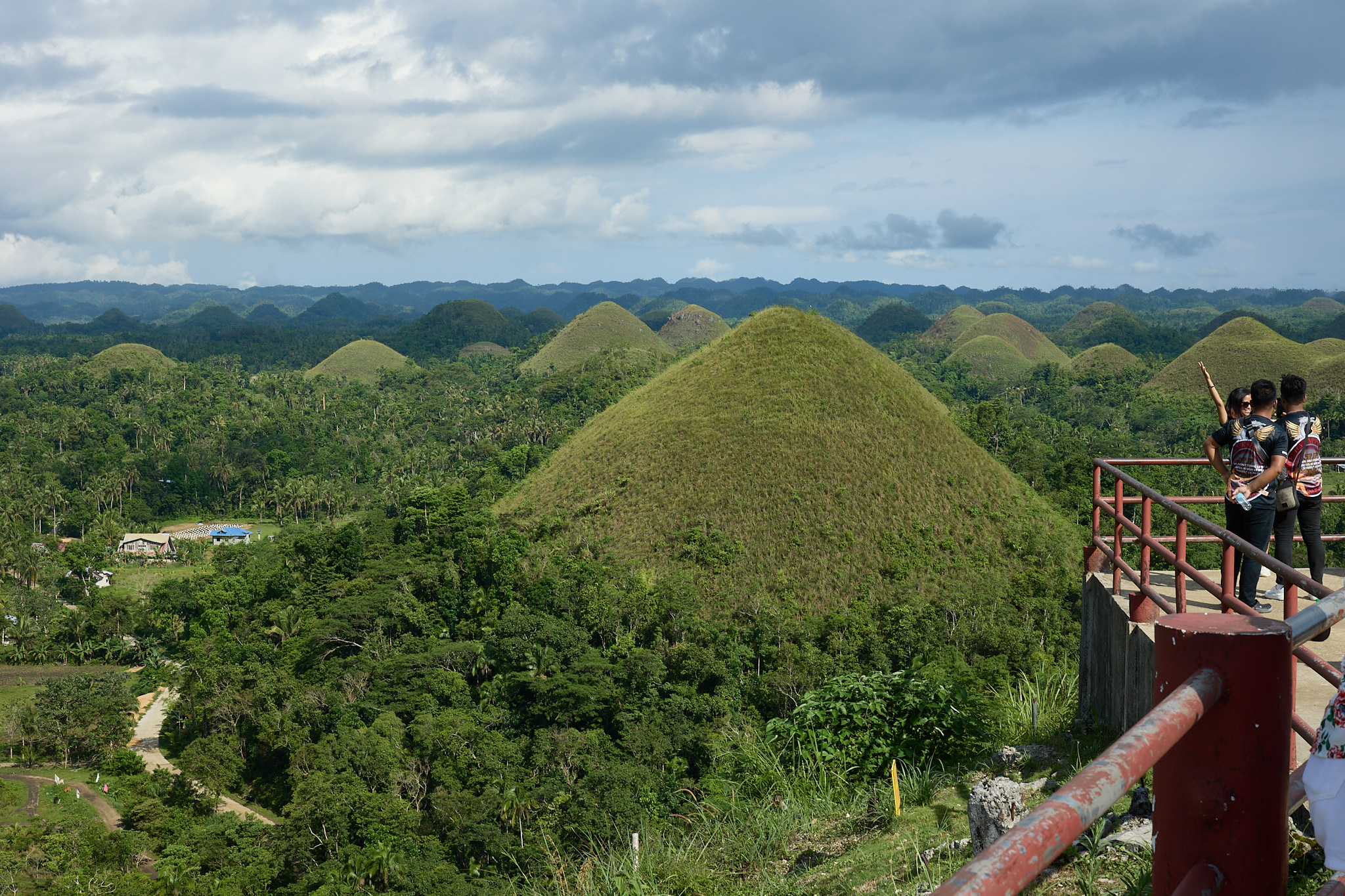 A grass-covered cone-shaped hill rising out of forest, with dozens of similar hills behind it and visitors at a red railing on the viewing deck to the right