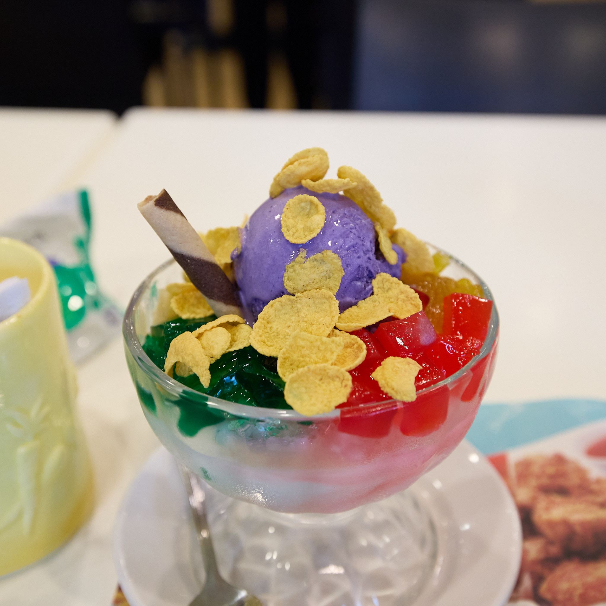 Halo-halo in a footed glass on a saucer: a scoop of purple ice cream scattered with cornflakes, green and red jelly-like pieces beneath, and a striped wafer roll stuck in at an angle.