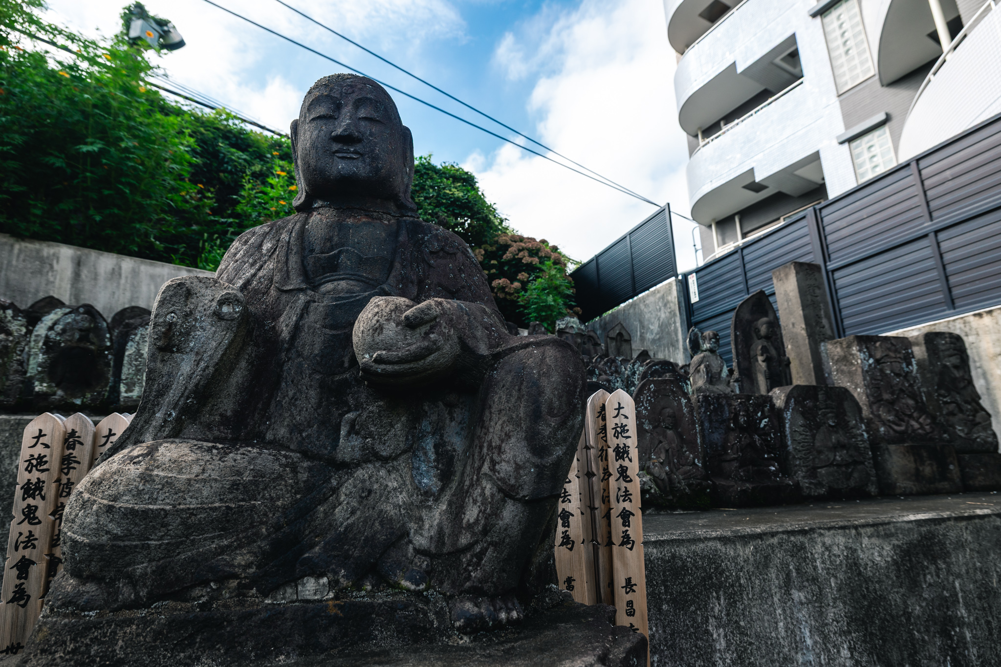 A seated figure in front of the smaller carved stones, with apartment balconies and utility wires beyond.