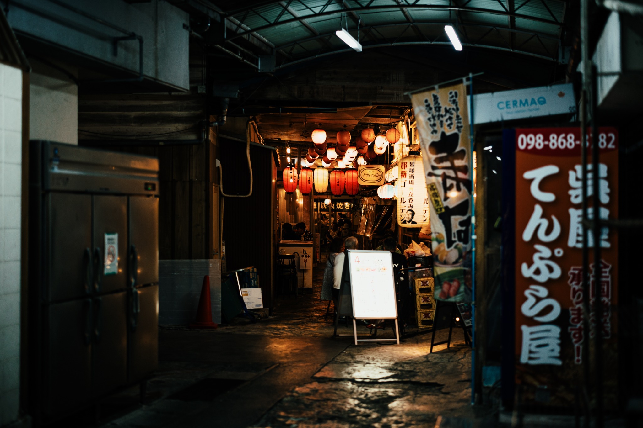 A dim covered passage leading to small eateries lit by red and white lanterns.
