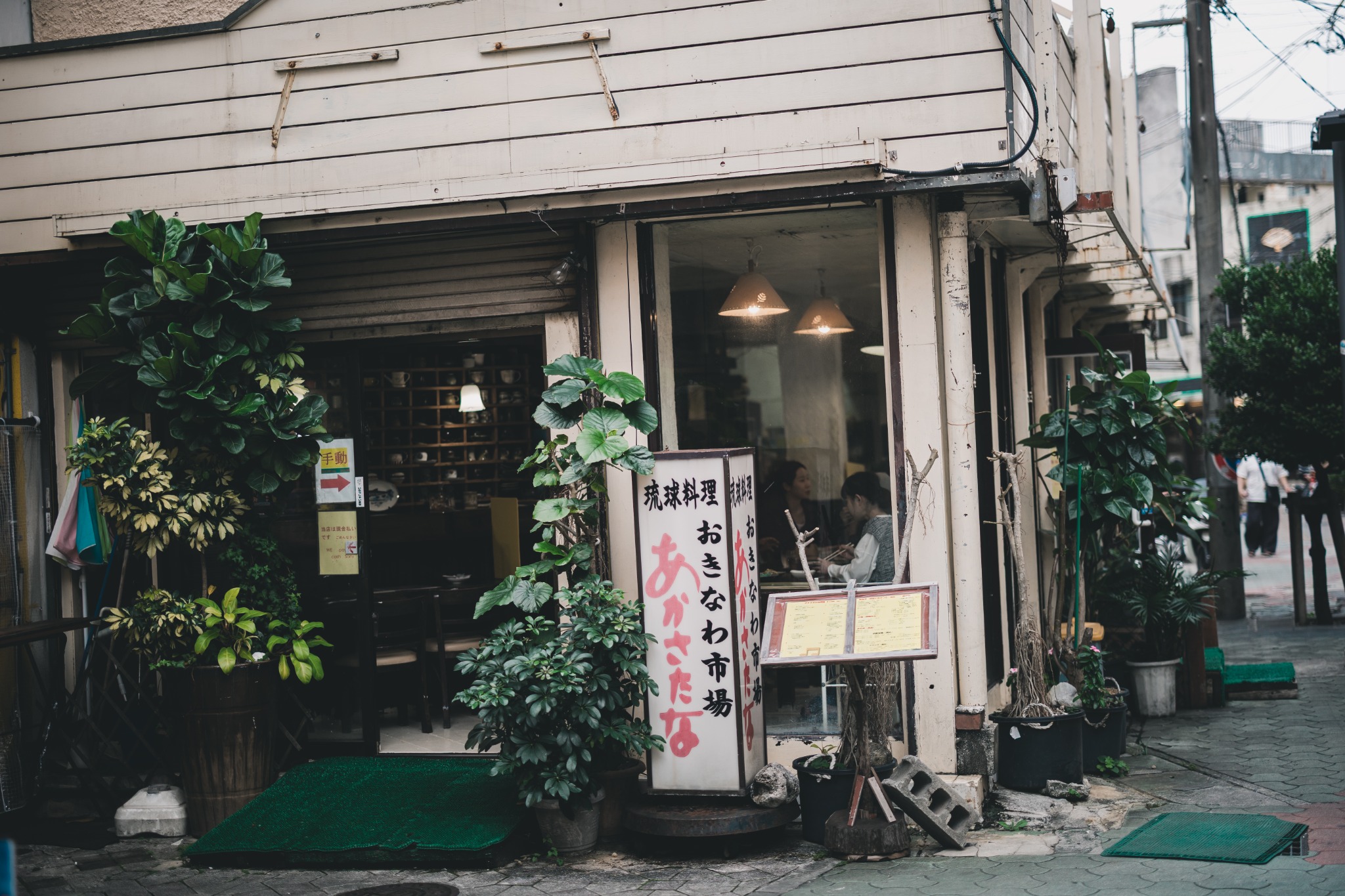 A plant-filled restaurant frontage with Japanese signs, an outdoor menu and diners visible through the window.