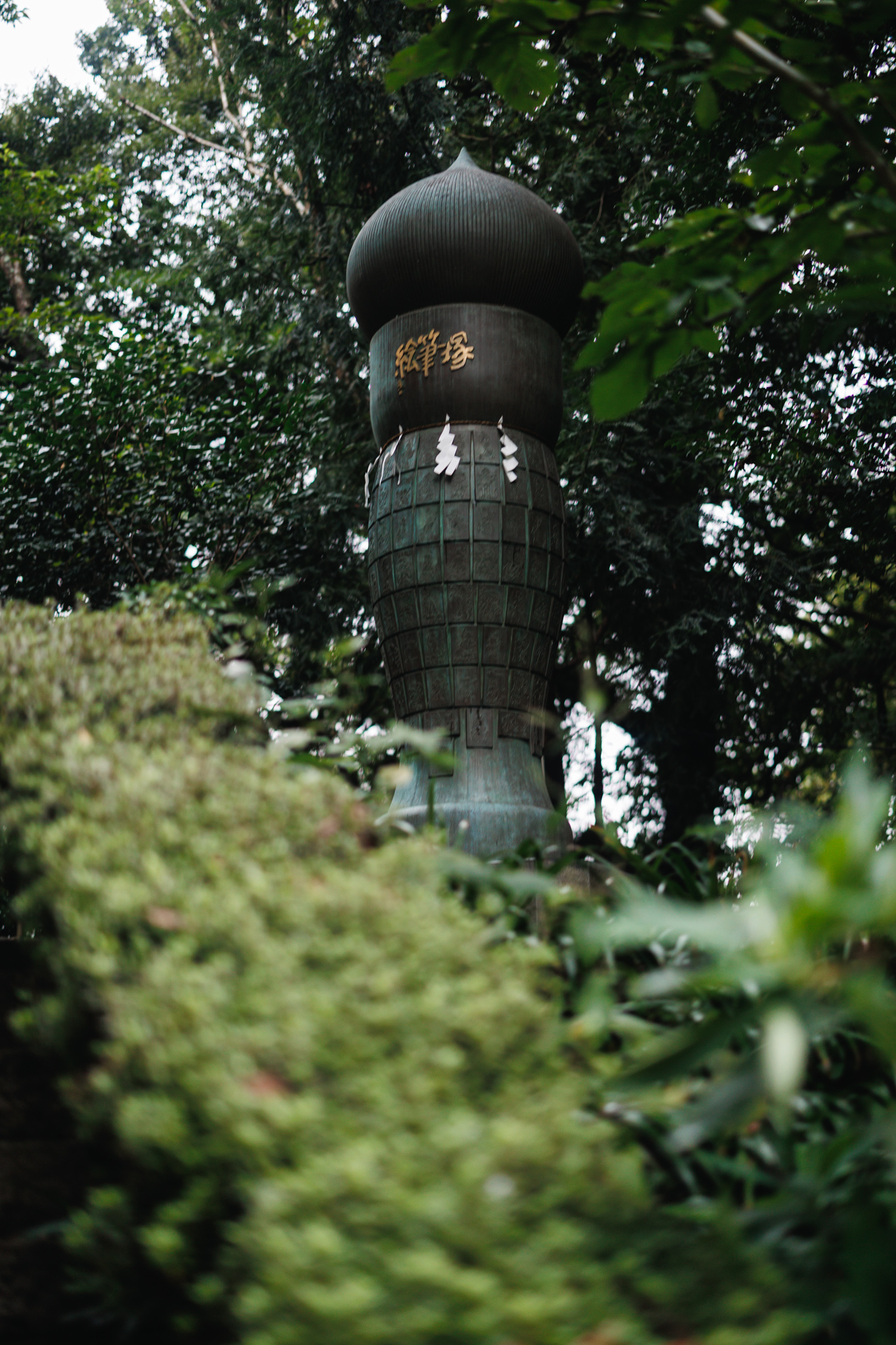 The bronze Efudezuka rising above shrubbery among trees at Egara Tenjin Shrine
