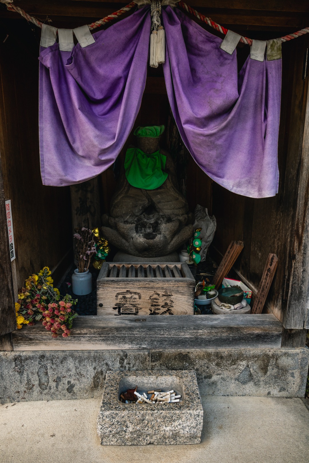 Enmei Tabako Jizō inside a small wooden shrine in Yudanaka, wearing green cloth beneath purple curtains, with flowers and cigarettes in a stone container in front.