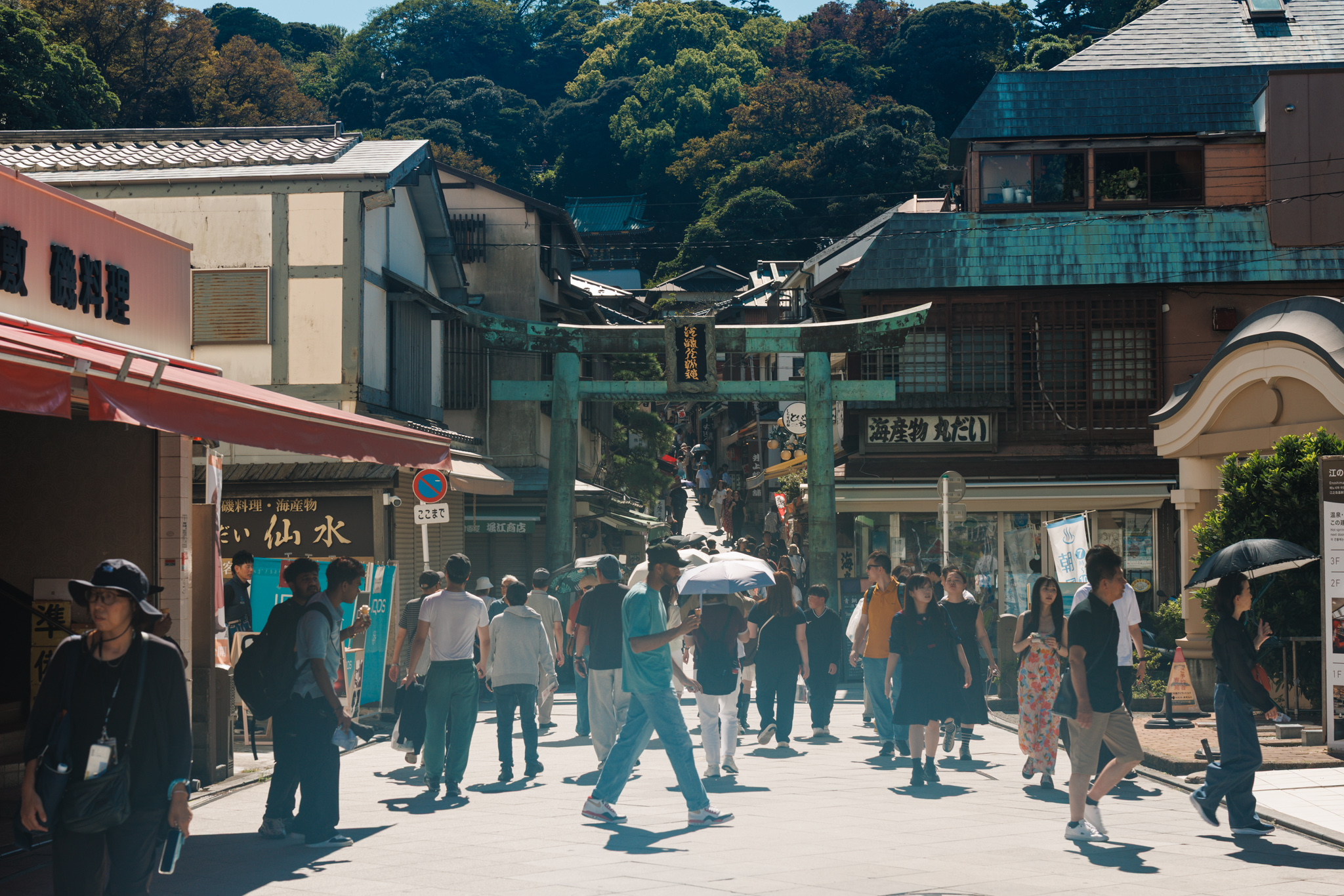 The green bronze torii at the foot of the shopping street leading to Enoshima Shrine, with a crowd of visitors passing beneath it and wooded hills behind