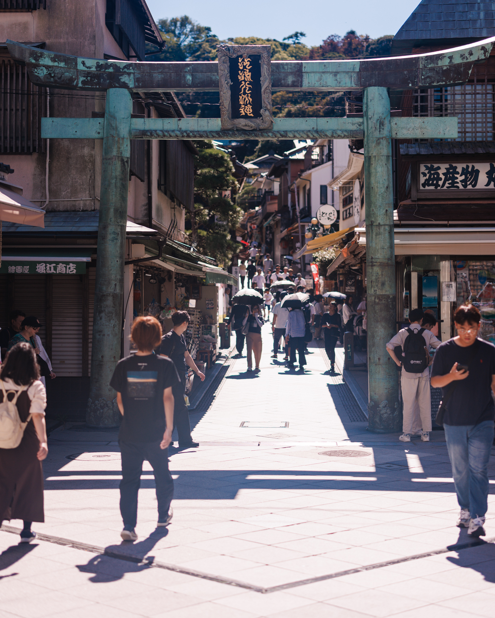 The bronze torii seen from the street, its patina flaking on the crossbeams and a cast-bronze sacred rope along the lower beam, with the narrow approach climbing beyond