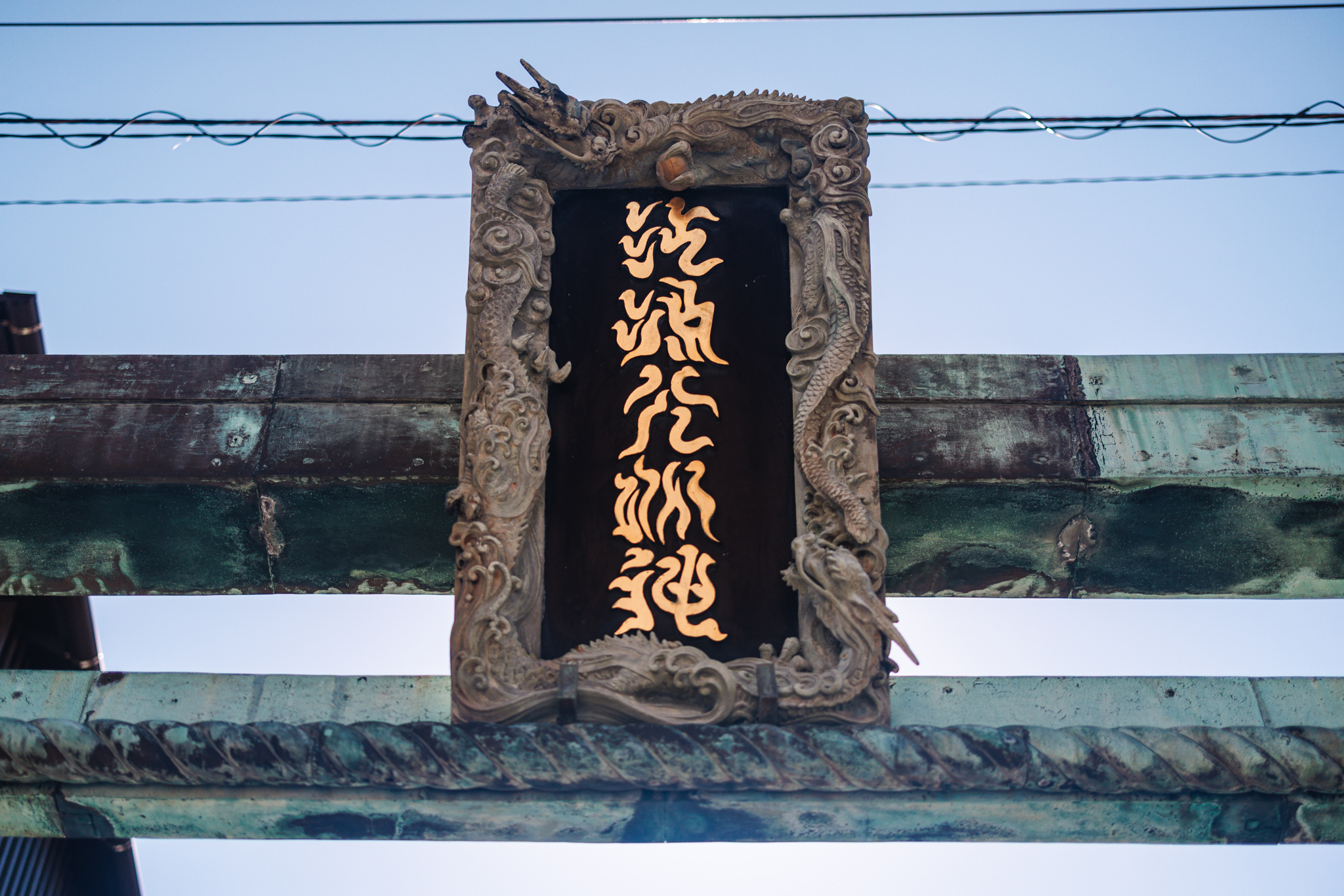 The torii’s plaque reading Enoshima Daimyōjin in gold, snake-like strokes, framed by carved dragons among clouds and waves, above the bronze rope on the crossbeam