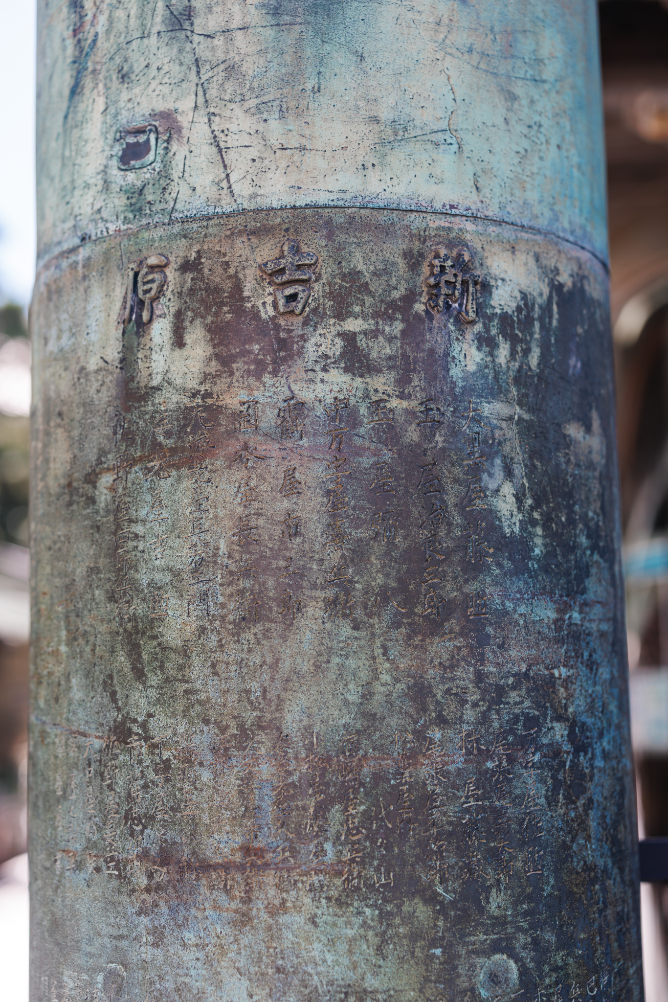 A weathered bronze pillar of the torii, with the characters 新吉原 (Shin-Yoshiwara) cast in relief above faint engraved columns of donors’ names
