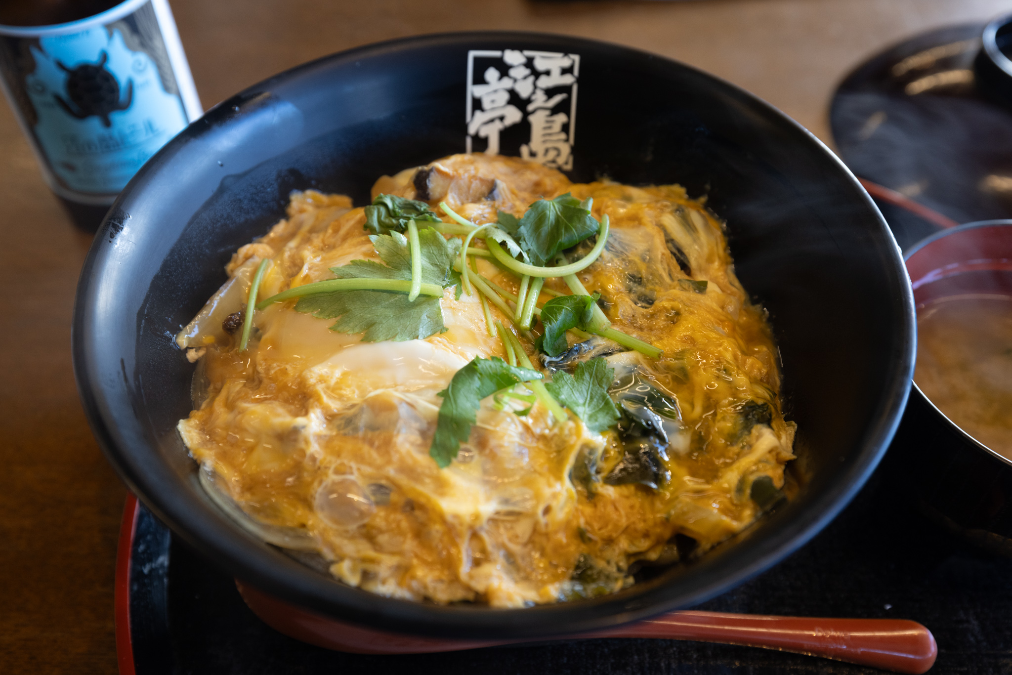 A black bowl of Enoshima-don: turban shell and onion bound in soft egg, topped with mitsuba, with the Enoshimatei logo on the bowl’s rim