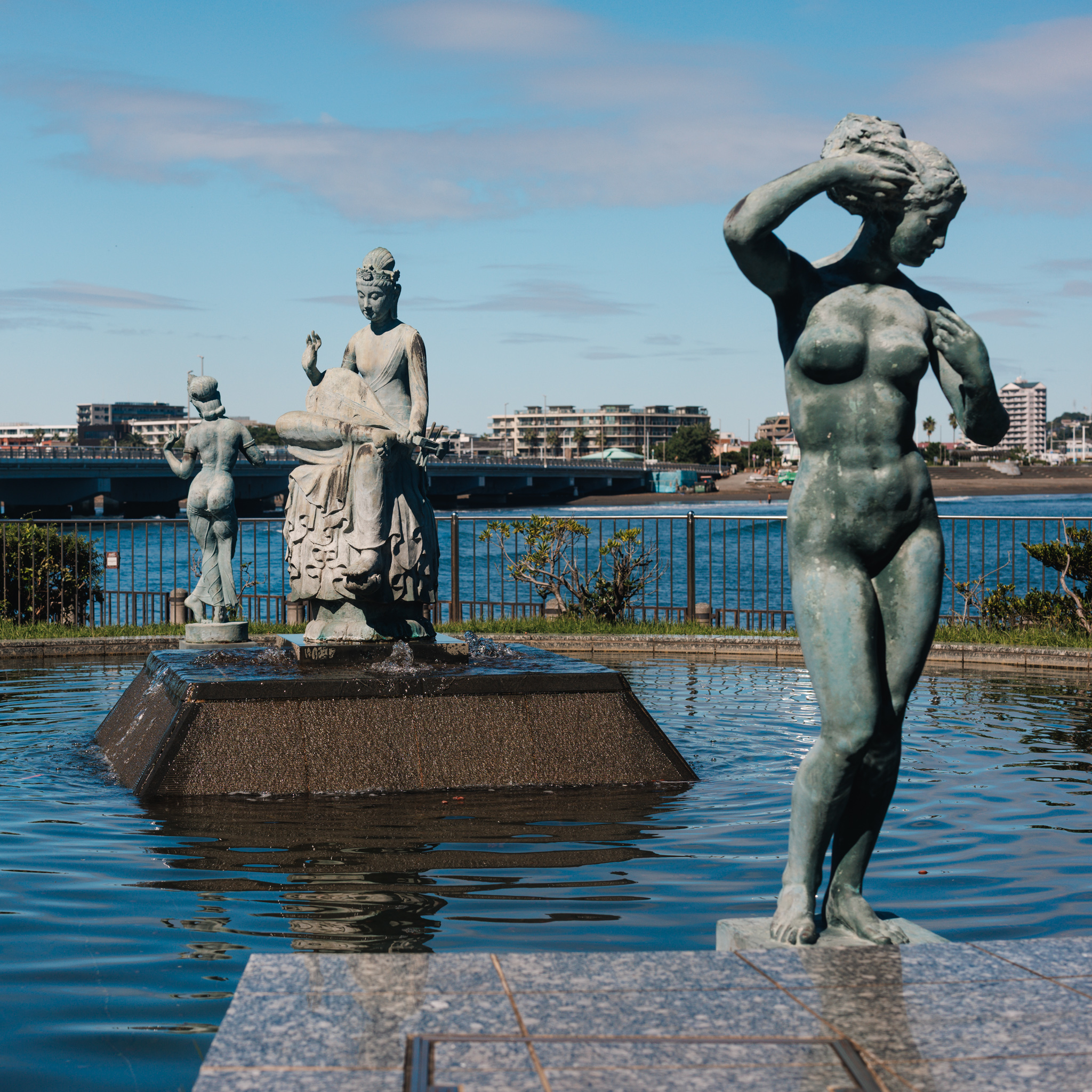 A bronze female nude at the edge of the Enoshima fountain, with Benzaiten playing a lute and a dancer behind her and the bridge to the mainland beyond