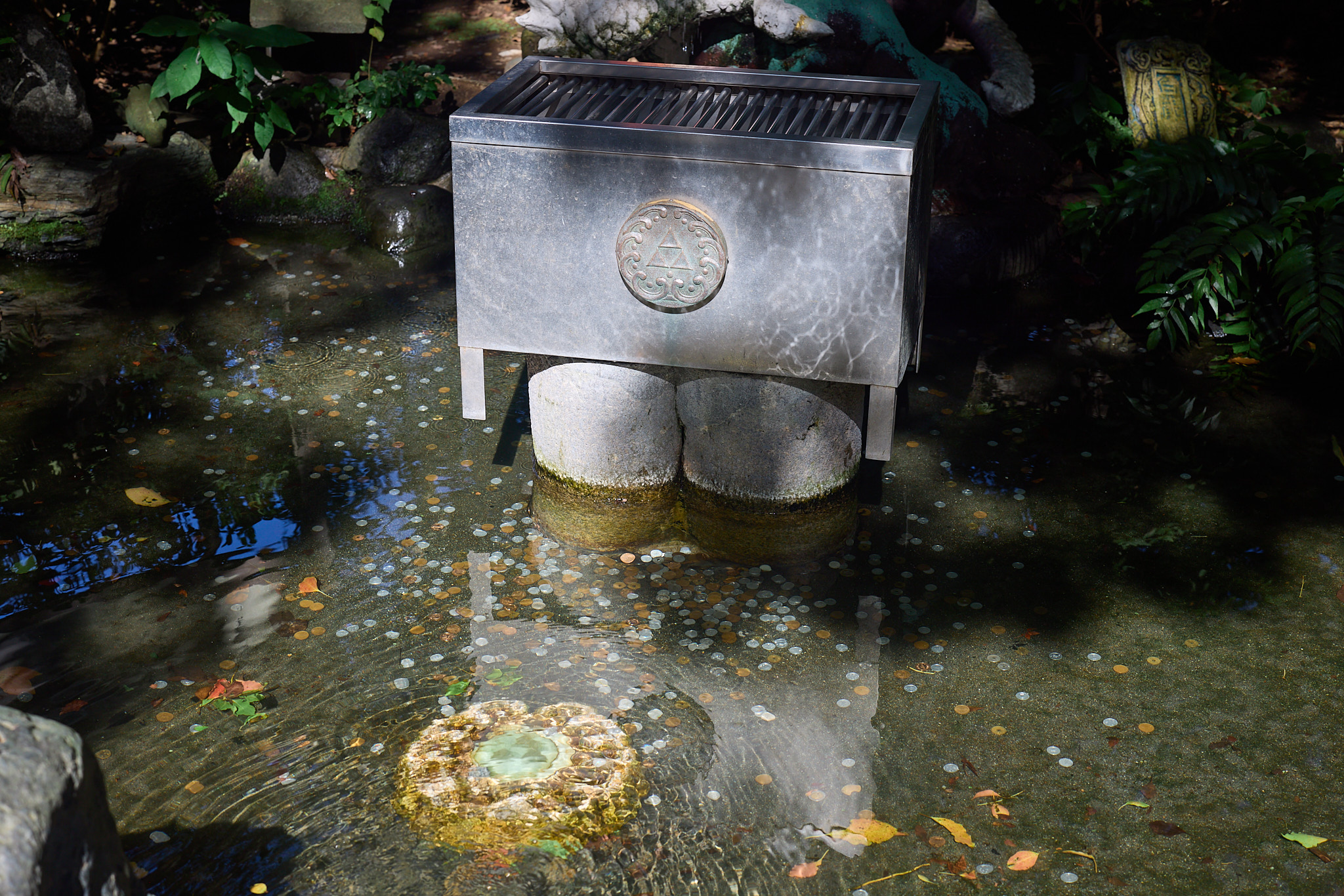 A metal offertory box on stone supports in a shallow pool whose bottom is covered with scattered coins