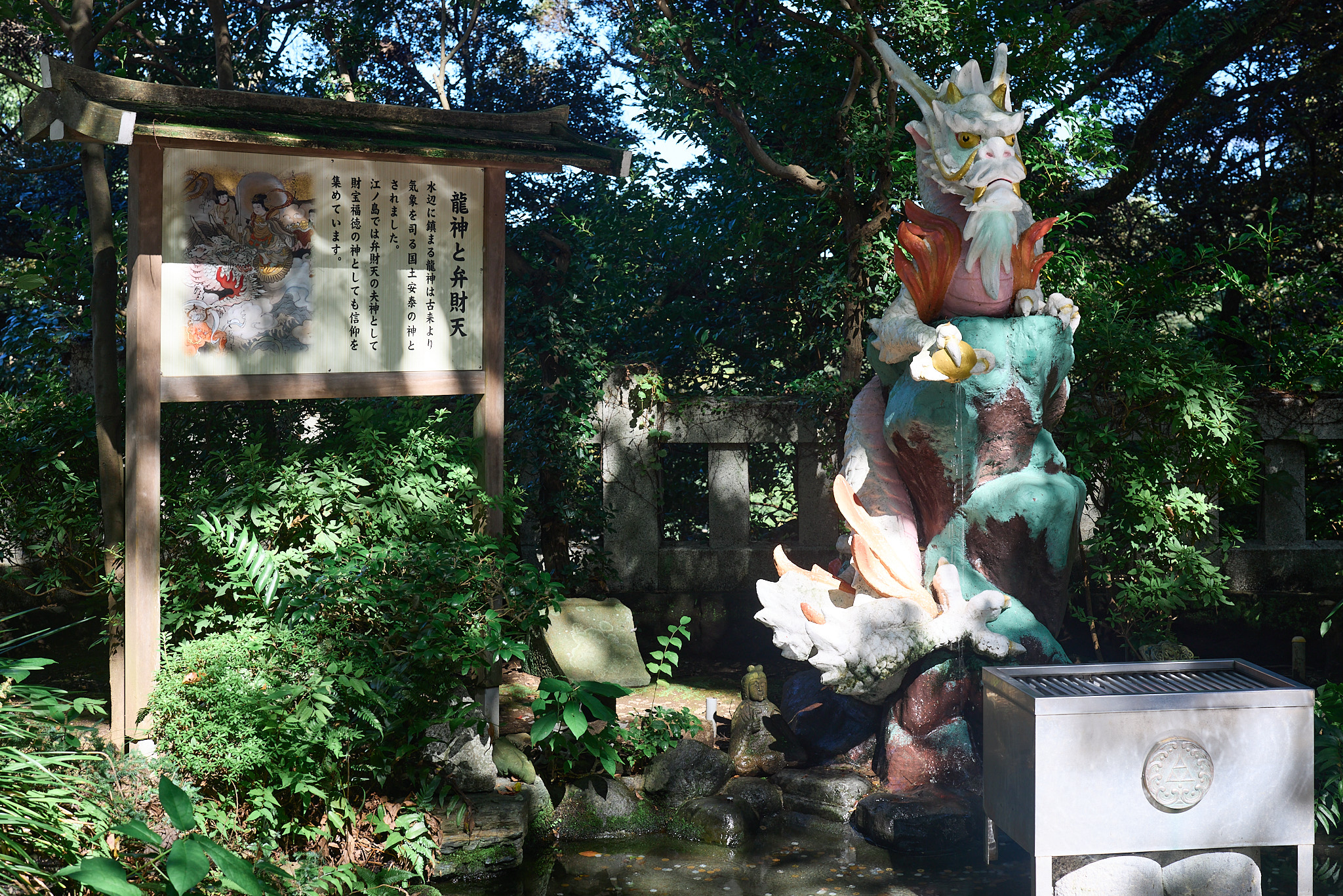 A roofed wooden signboard with a painting of a goddess and a dragon and vertical Japanese text, beside a white dragon statue above a pool among trees