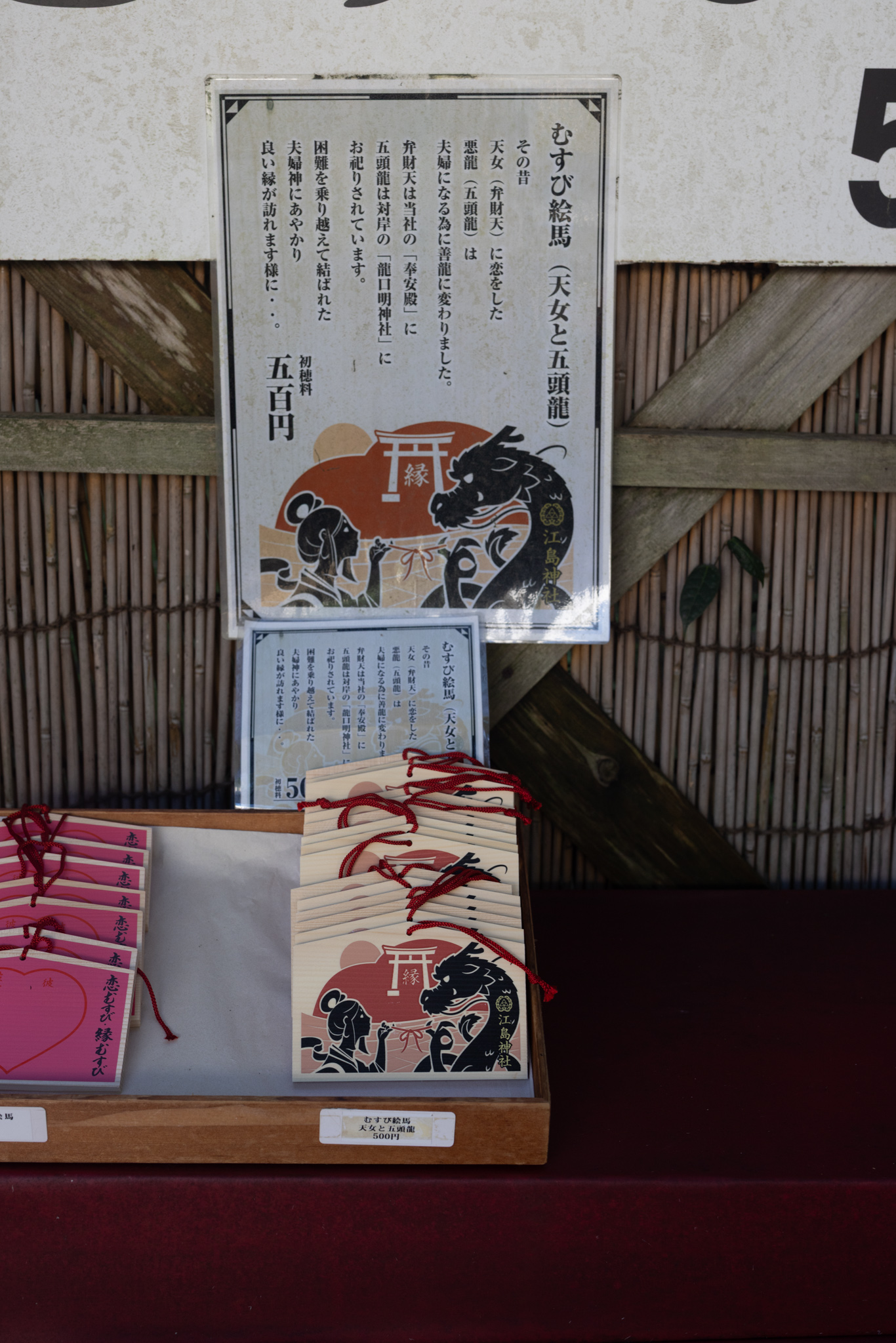 A printed sign showing a woman in silhouette and a black dragon tied by a red thread under a red torii, with Japanese text above and wooden plaques with red cords below