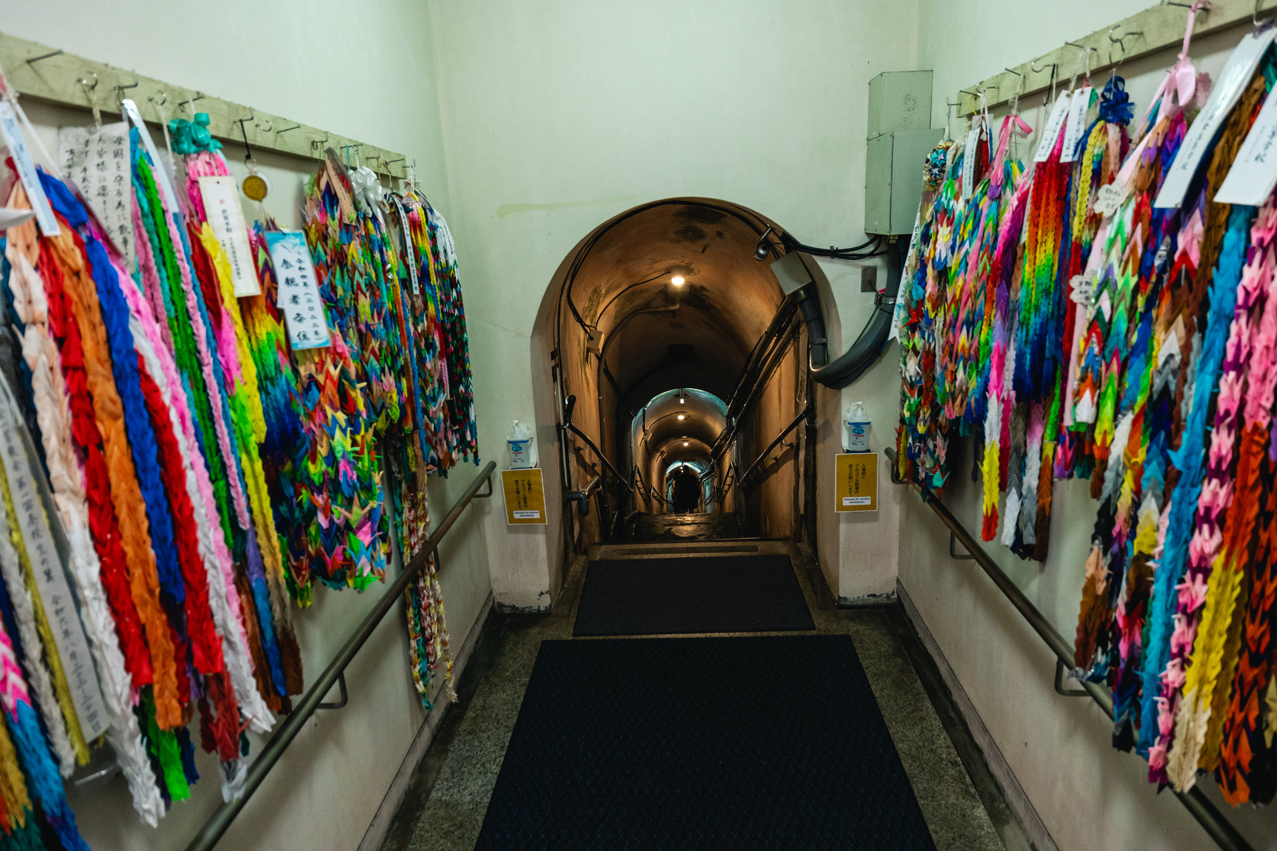 A corridor lined on both sides with long strings of brightly coloured paper cranes, leading to an arched opening where a lit staircase goes down into the tunnels