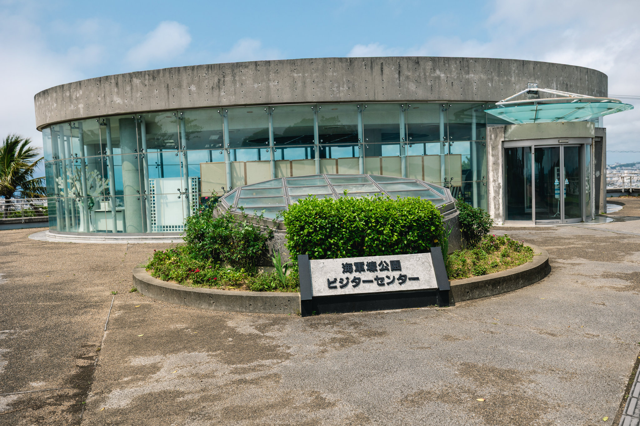A round concrete building with curved glass walls on a hilltop, with a glazed dome among shrubs and a sign reading 海軍壕公園 ビジターセンター
