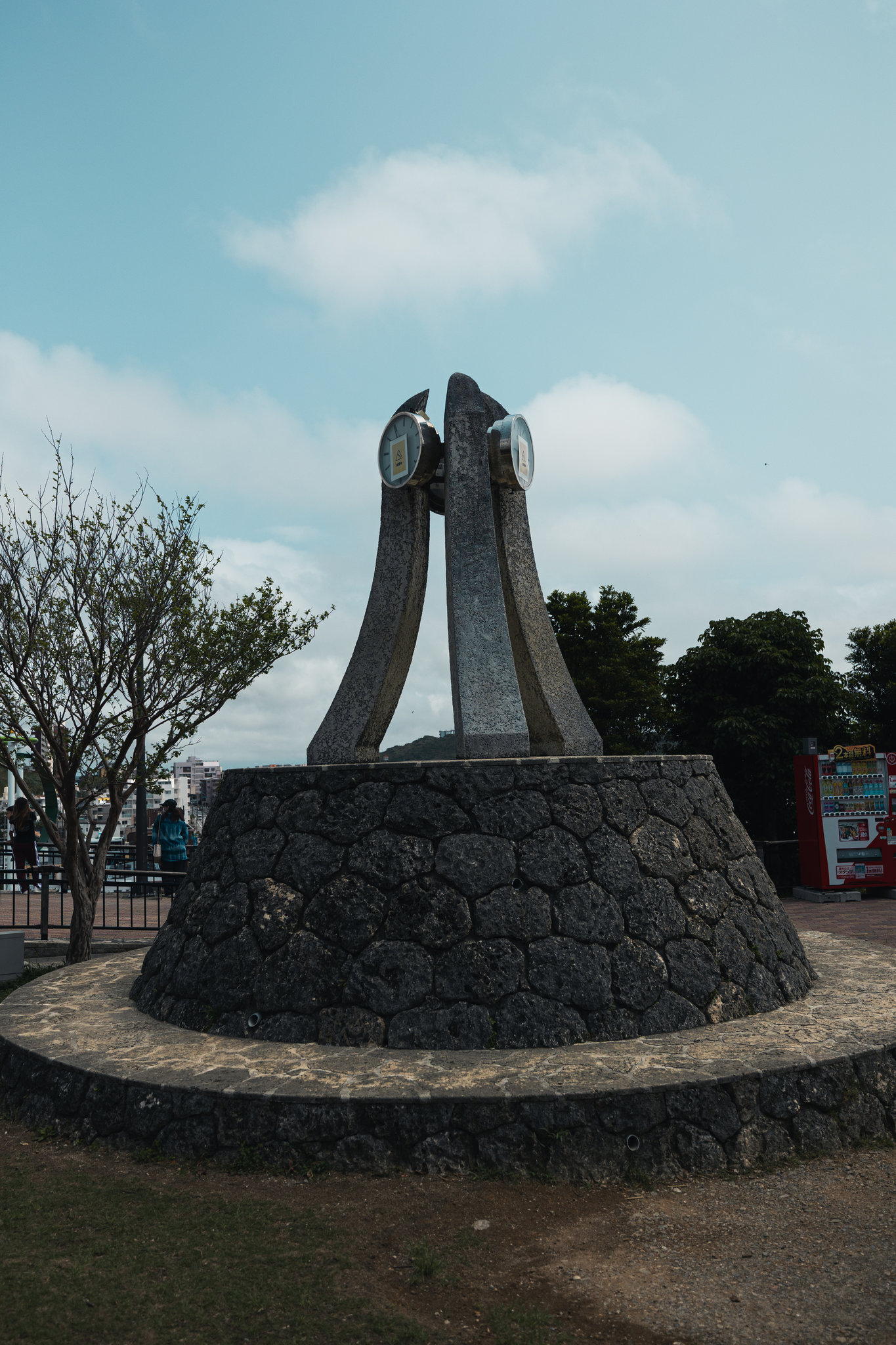 A clock tower of three tall curved stone fins on a round base of dark coral-stone masonry, its two clock faces covered with paper notices, under a cloudy sky
