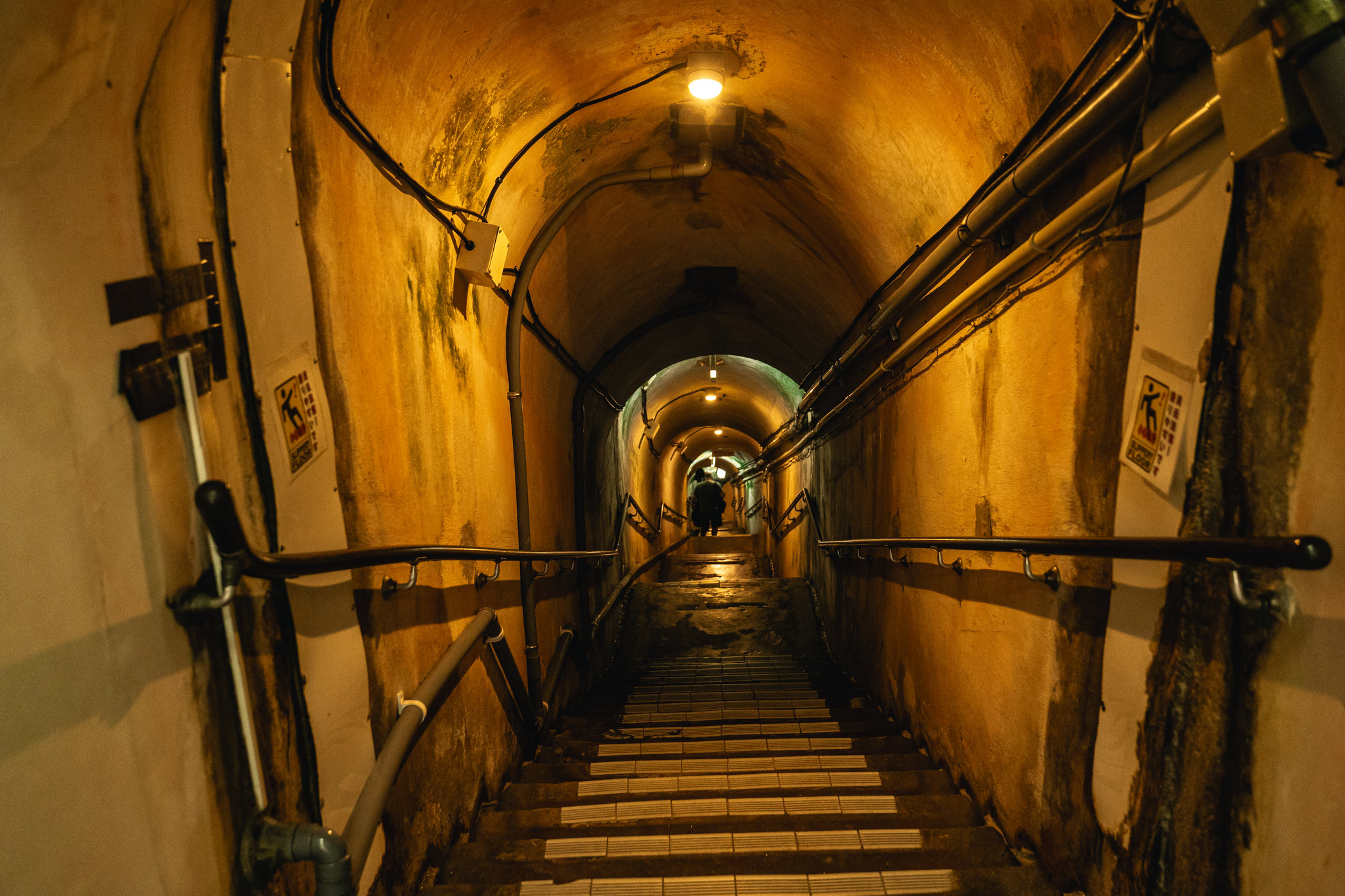 A steep staircase descending through a vaulted tunnel with yellowed walls, handrails and cables, lit by ceiling lamps, with a person far down the steps