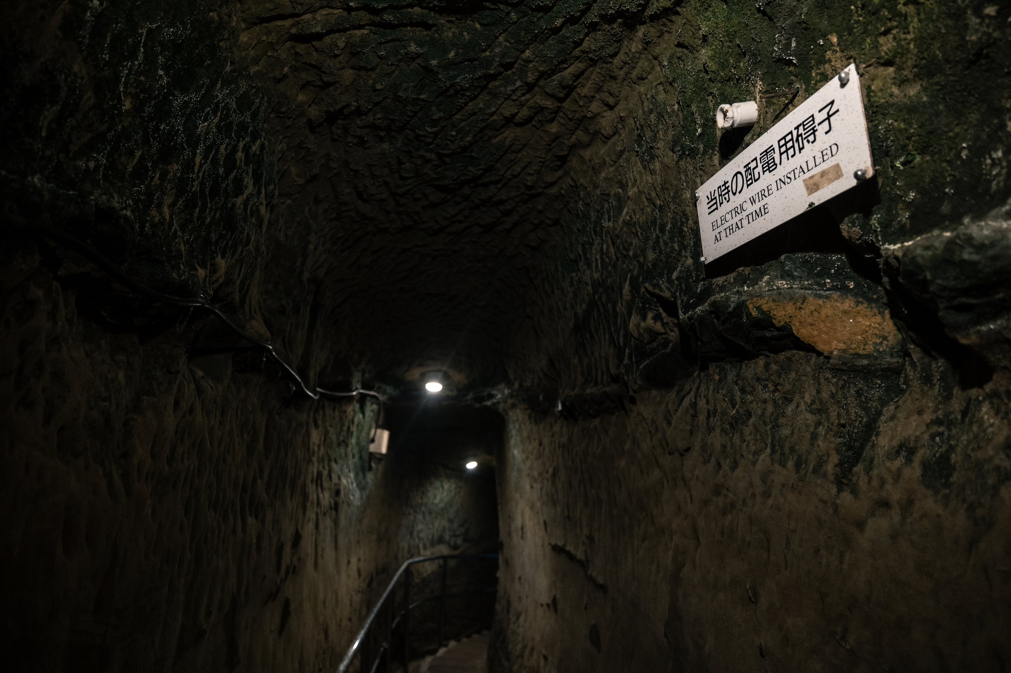 A narrow, dark tunnel cut through rough rock, with a small white insulator high on the wall above a sign reading 当時の配電用碍子 and “Electric wire installed at that time”