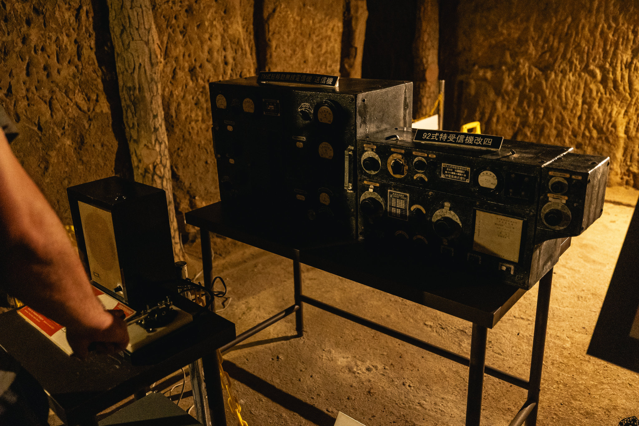 Two black radio sets with dials and meters on a metal table against a rock wall, labelled as a transmitter and a Type 92 receiver, with a visitor’s arm reaching to a telegraph key at the left